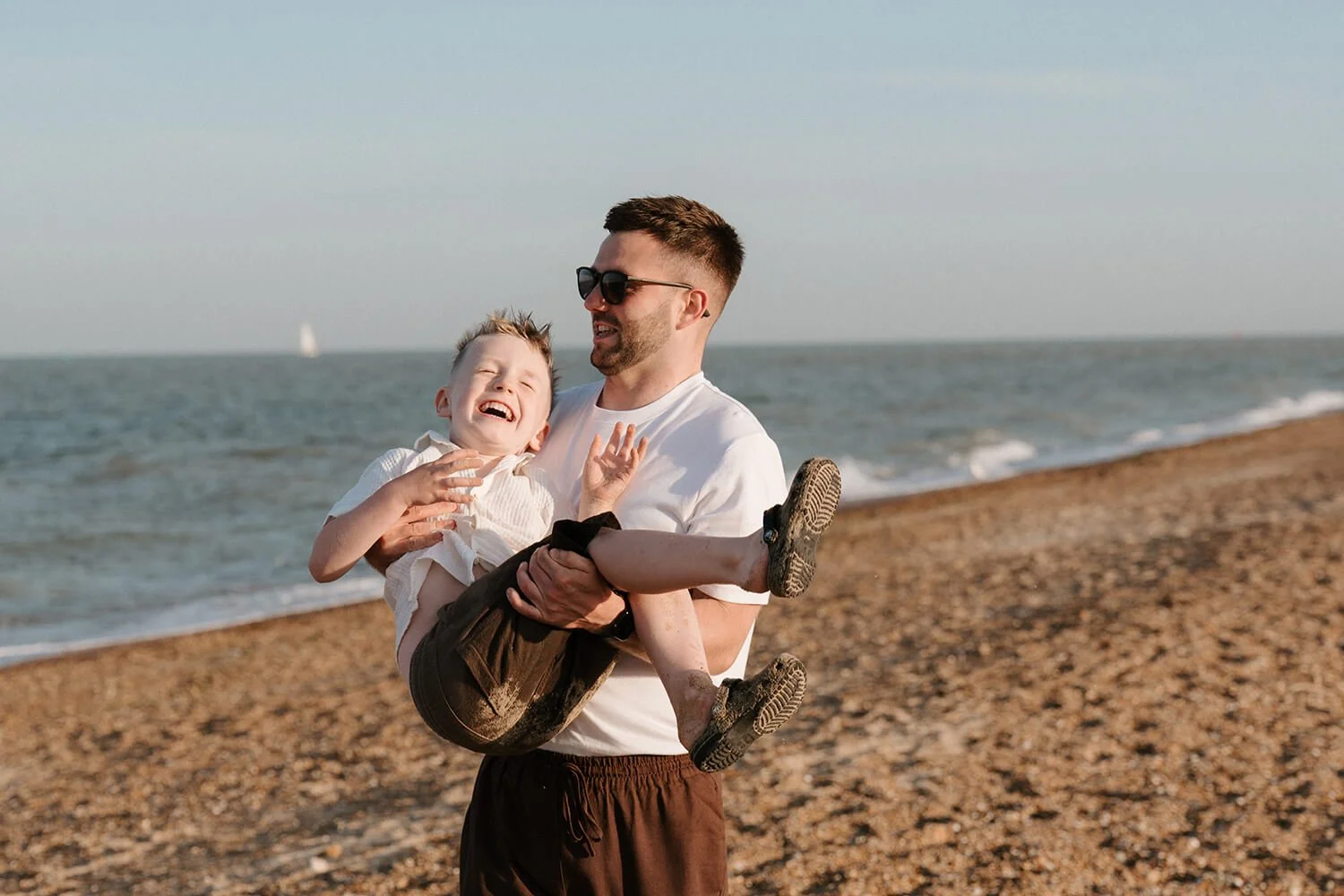 A man holding a laughing boy on a pebble beach by the ocean with sailboats in the background.