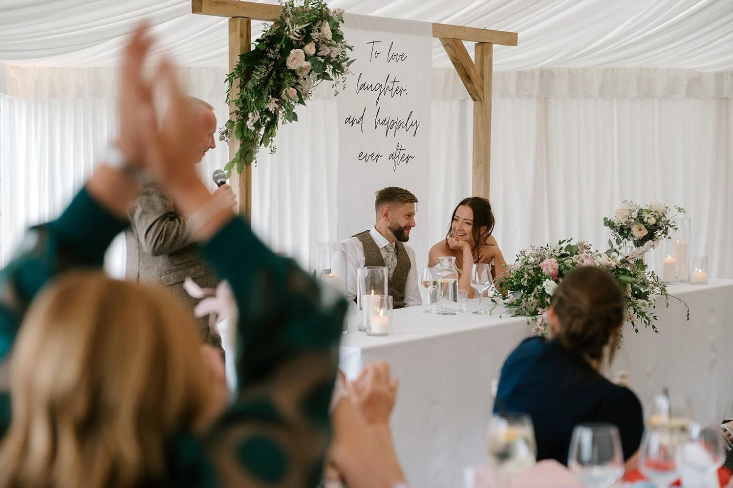 A wedding reception with a bride and groom sitting at a decorated table, smiling at each other, with a man giving a speech in front, and guests raising glasses.