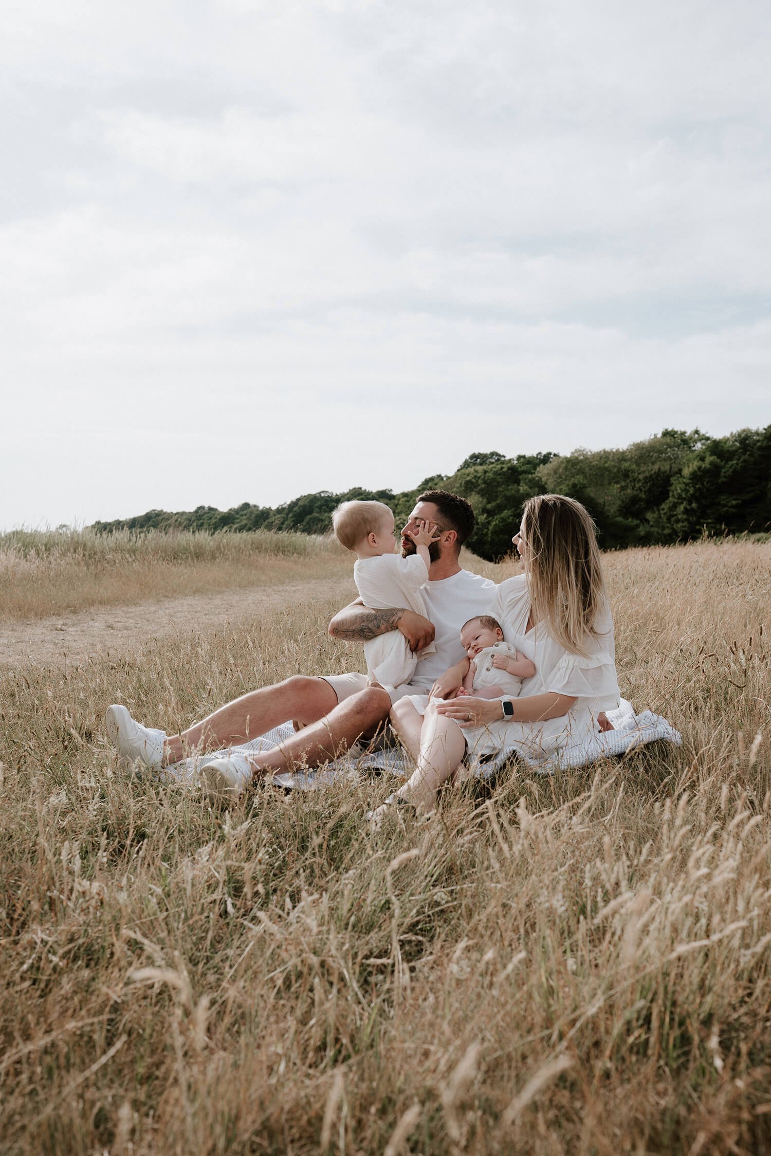 A family of four sitting on a blanket in a grassy field, enjoying a moment together. The father, with a beard and tattoos, holds a young child who is touching his face. The mother, with blonde hair, holds a baby in her lap. The scene takes place outd