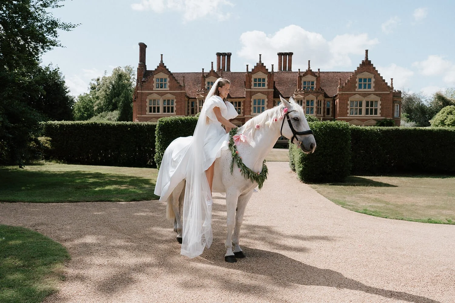 Bride in wedding dress riding a white horse decorated with pink flowers and greenery in front of a large red brick mansion.