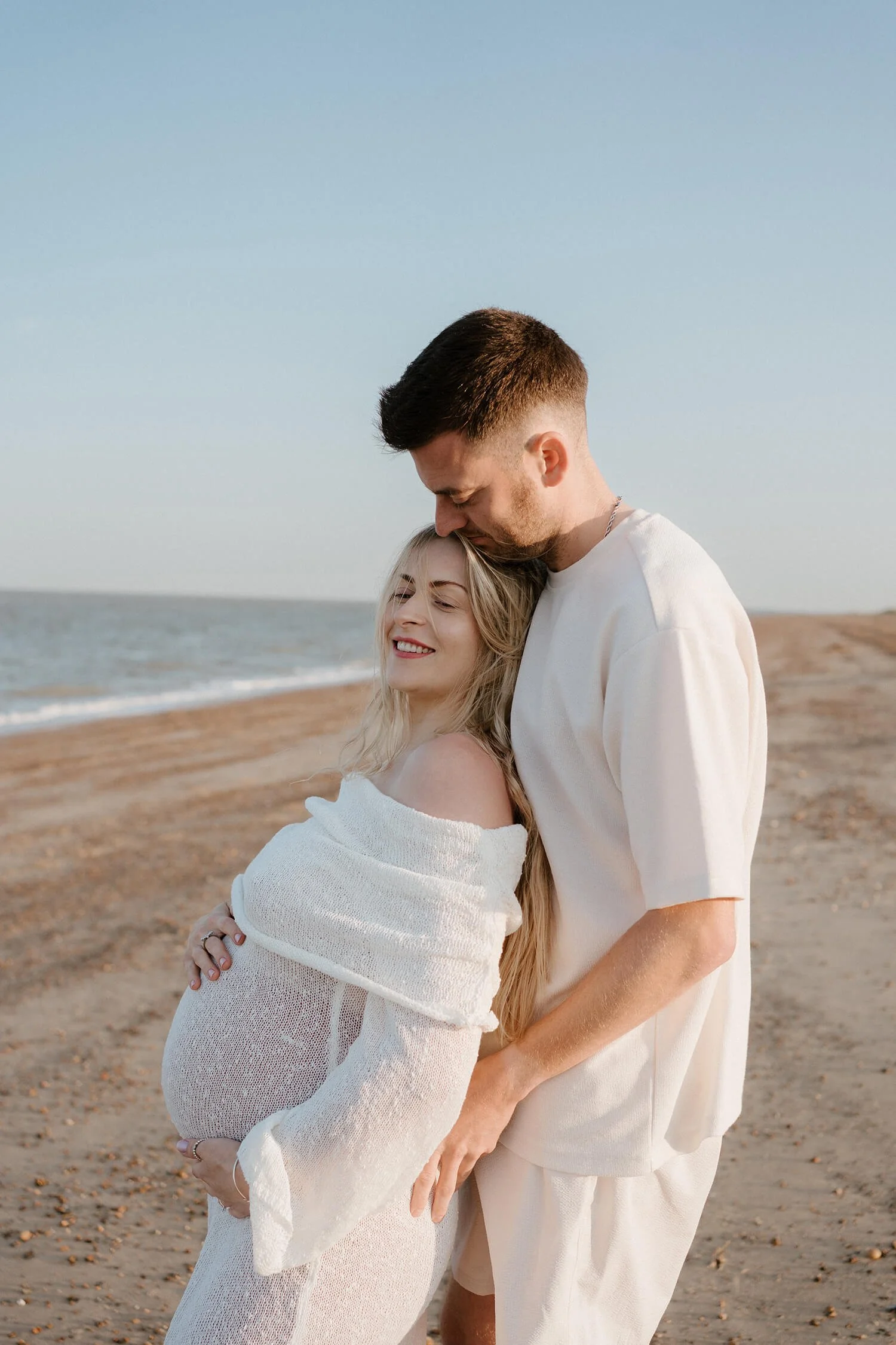 A pregnant woman in a white dress and a man in a white shirt on the beach, embracing and smiling with the ocean in the background.