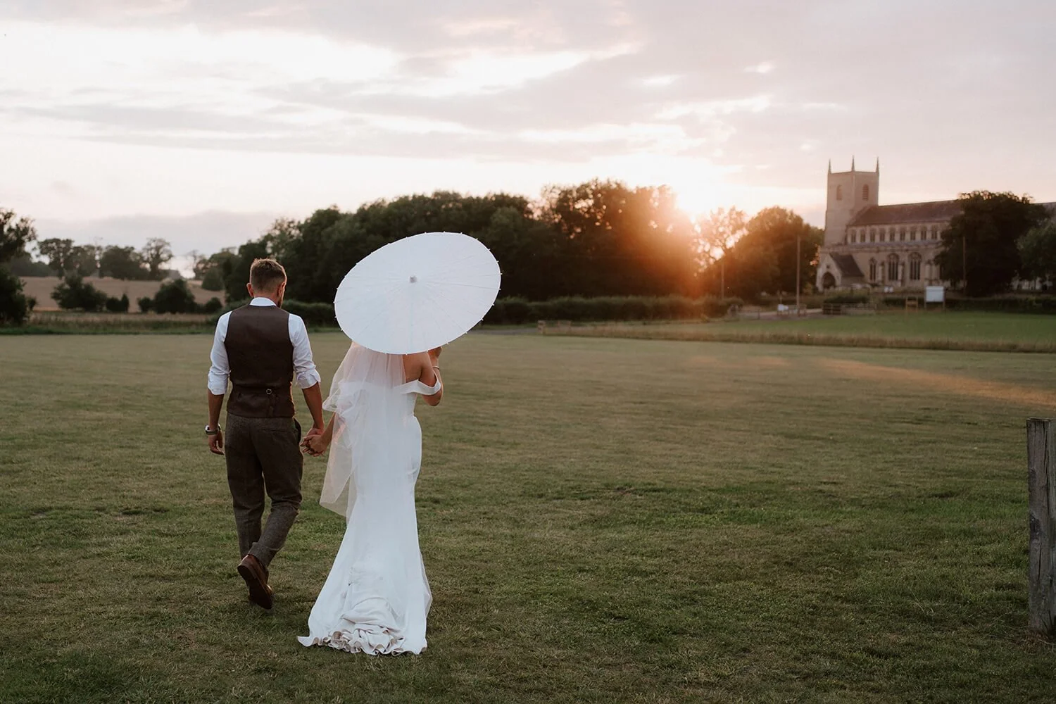 A bride and groom walking hand in hand across a grassy field at sunset, with a church in the background.