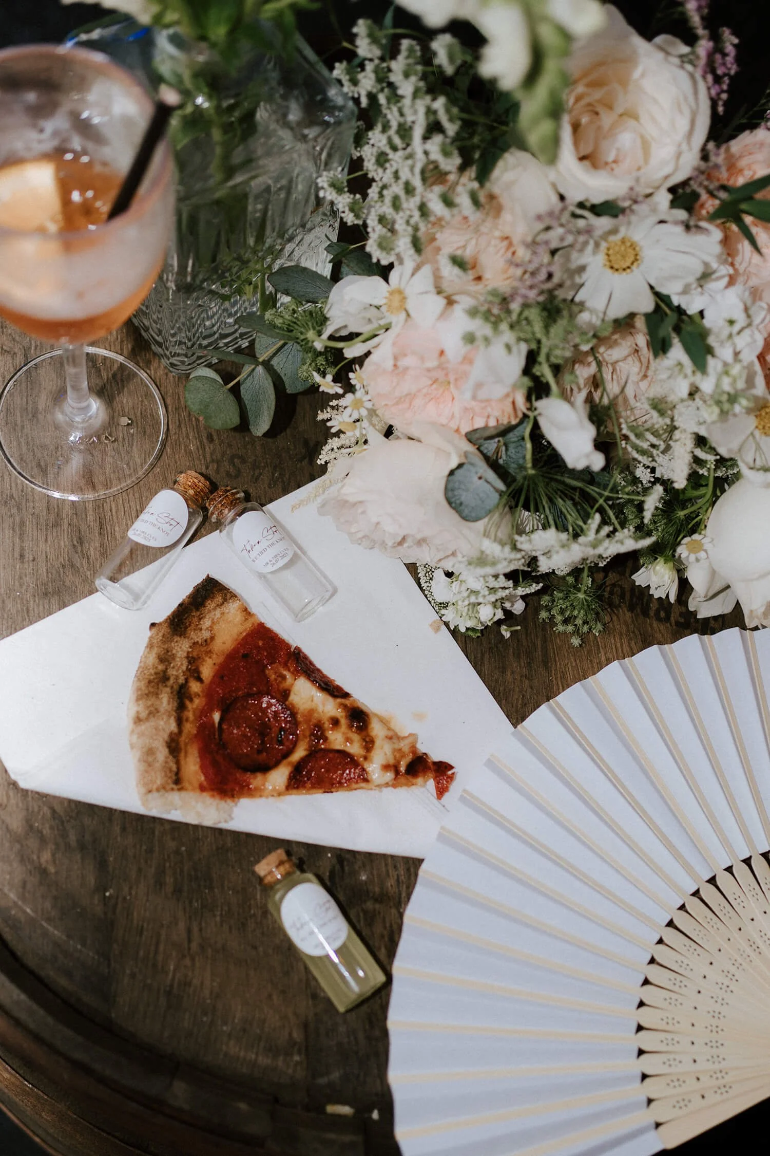 A slice of pepperoni pizza on a white paper plate, a glass of iced drink with lemon, small bottles of essential oils, and a large bouquet of white and pink flowers on a wooden table, with a white folding fan partially visible.