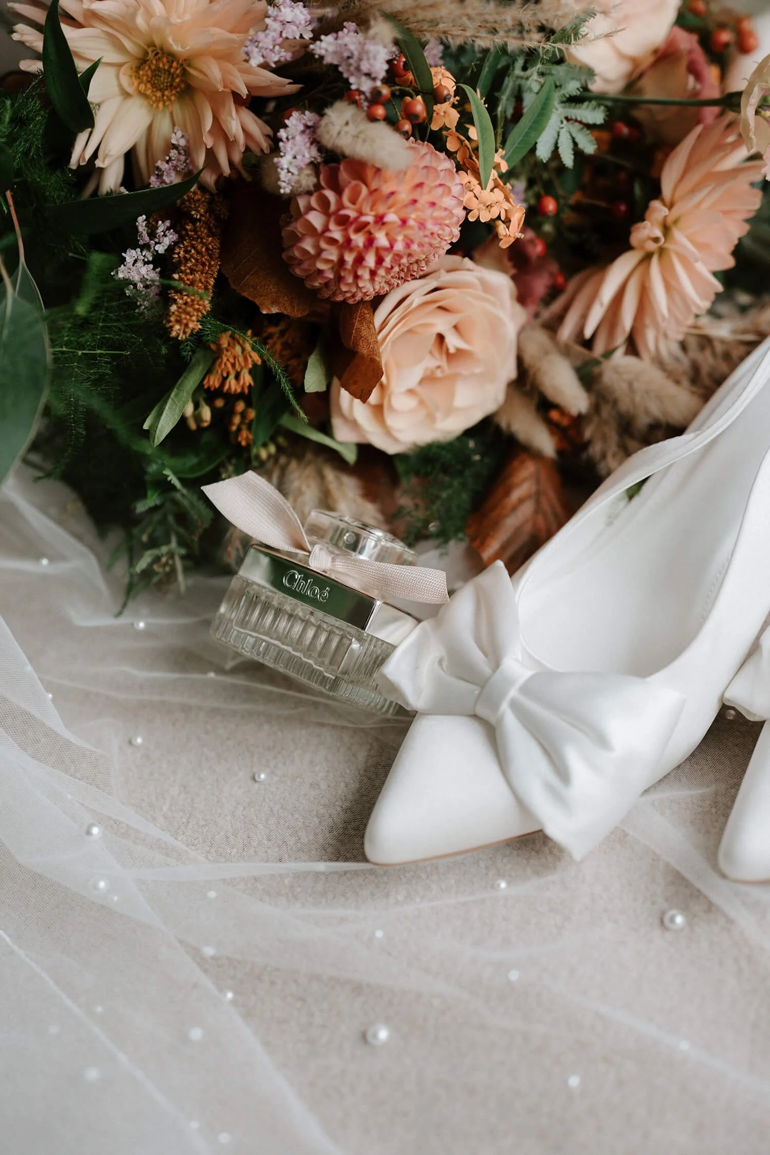 A close-up of a bouquet of flowers including roses and other blooms, a white high-heeled shoe with a bow, a perfume bottle labeled Chloé, and a sheer fabric with pearl accents.