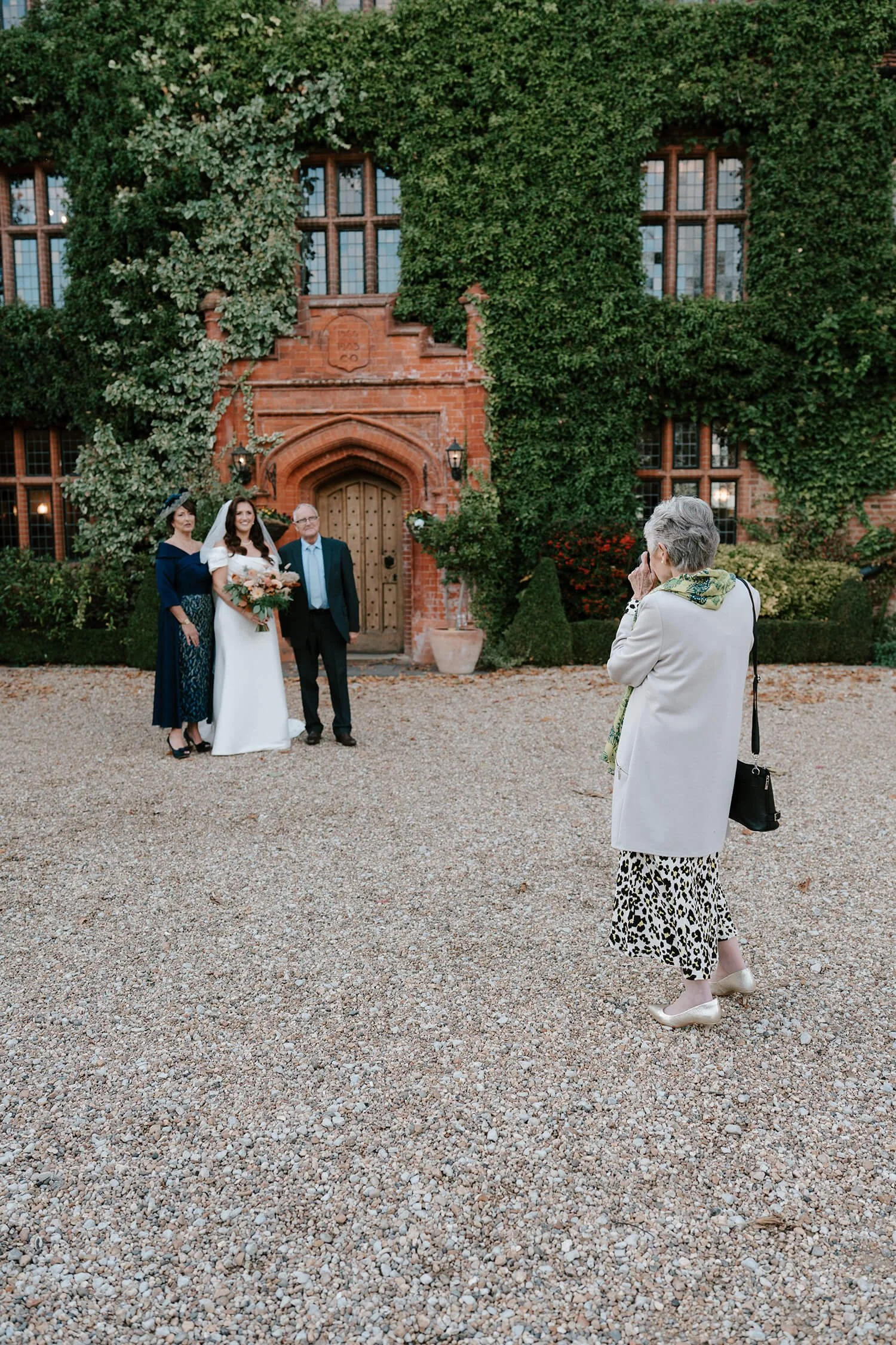 A bride and two older women standing in front of a red brick building with ivy, being photographed by an older woman in a white coat and leopard print skirt.