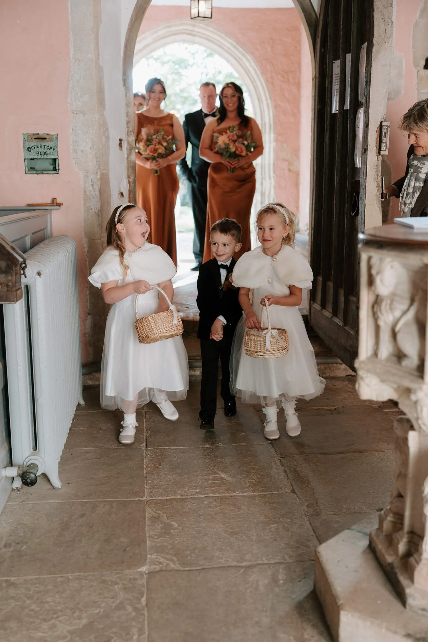 Three children, two girls in white dresses and one boy in a black suit, walking inside a church, holding baskets, with two women in brown dresses and a man in a tuxedo standing in the background at the church entrance.