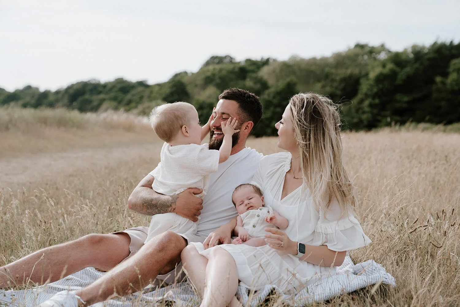 A happy family of four sitting on a blanket in a grassy field, with an adult male, adult female, and two young children, one of whom is touching the man's face.