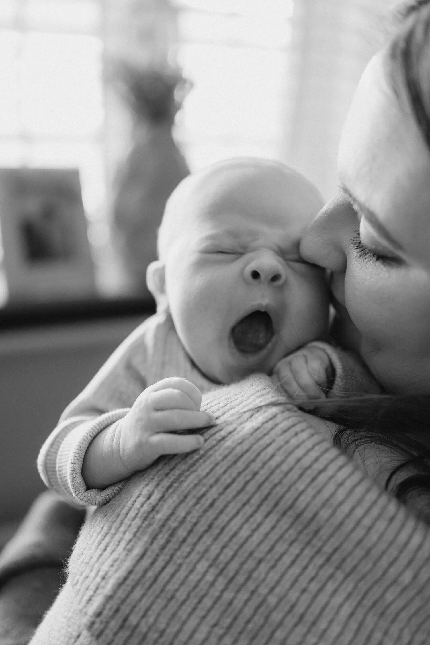 A woman holds a yawning newborn baby close to her face. The baby, with closed eyes and mouth open, is resting on the woman's shoulder. The image is in black and white.