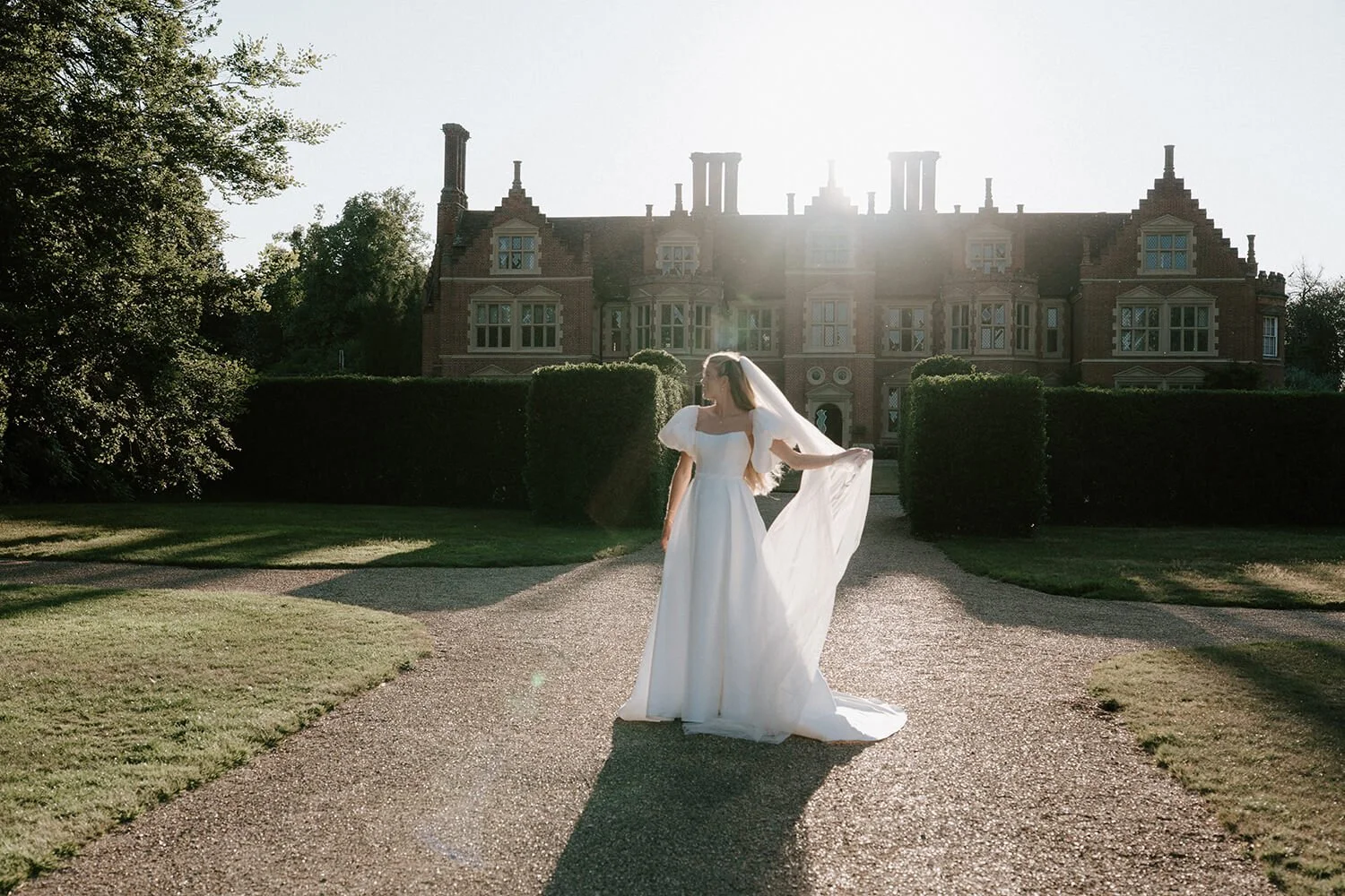 Bride in a white wedding dress standing on a gravel path in front of a large, historic brick mansion with lush greenery around, backlit by the setting sun.