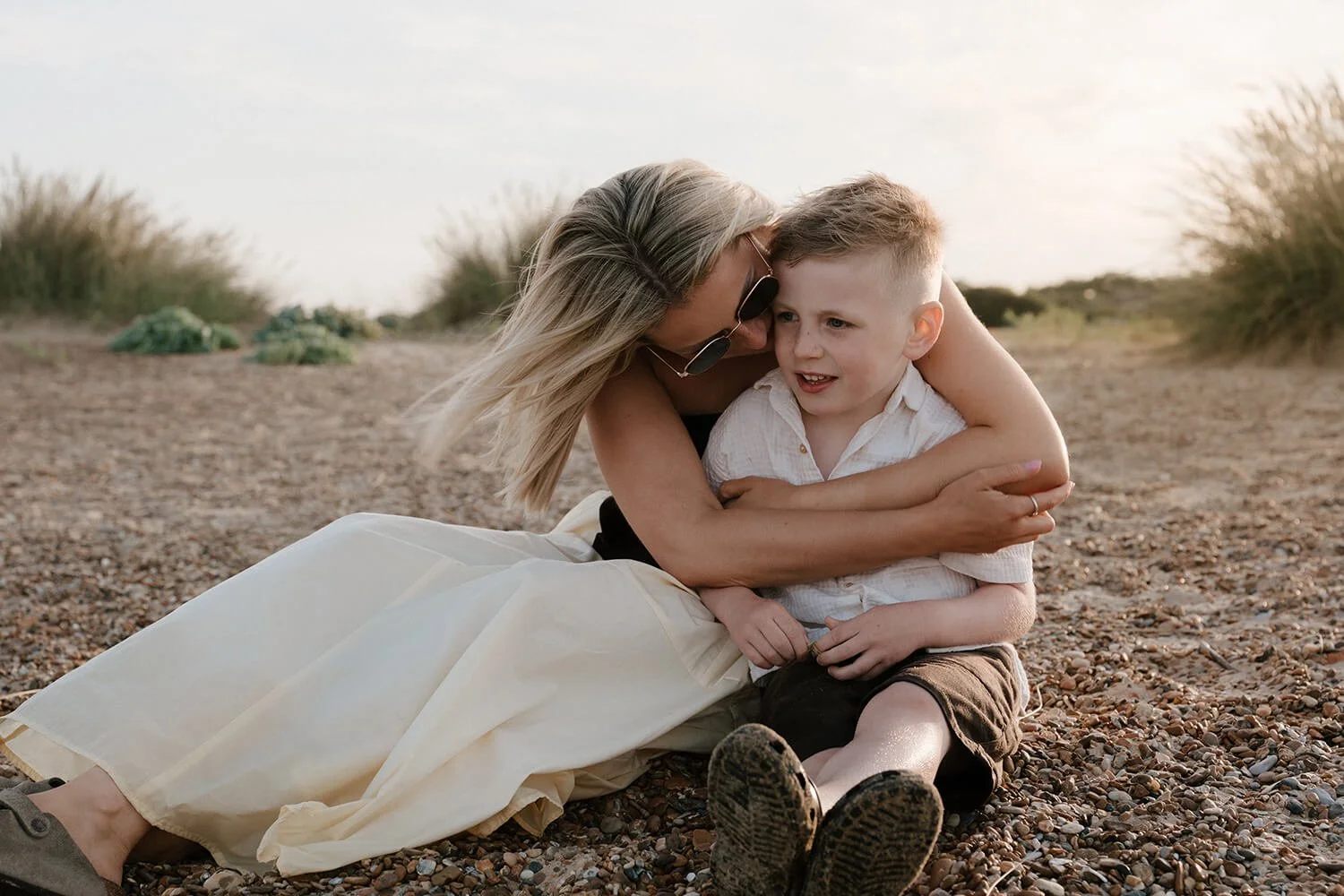 A woman and a young boy sitting on a pebble beach with tall grass in the background, embracing and talking during sunset.
