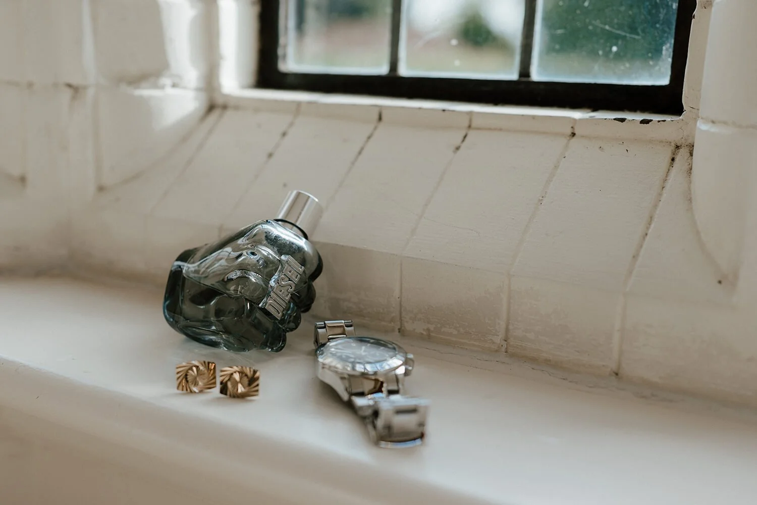 A pair of gold square-shaped earrings, a wristwatch with a metal band, a clear glass perfume bottle with a silver cap and black accents, all placed on a white windowsill next to a brown brick wall and a window with black frames.