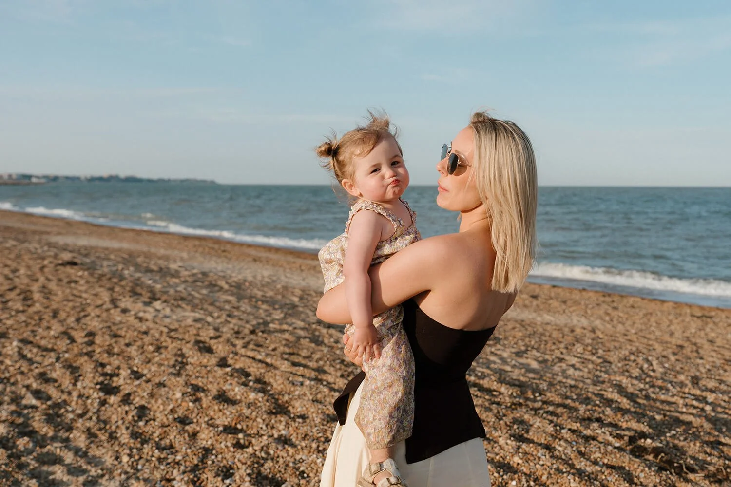 A woman with blonde hair wearing sunglasses holding a small girl with light brown hair styled in two buns on a pebble beach near the ocean, with a cloudy sky in the background.