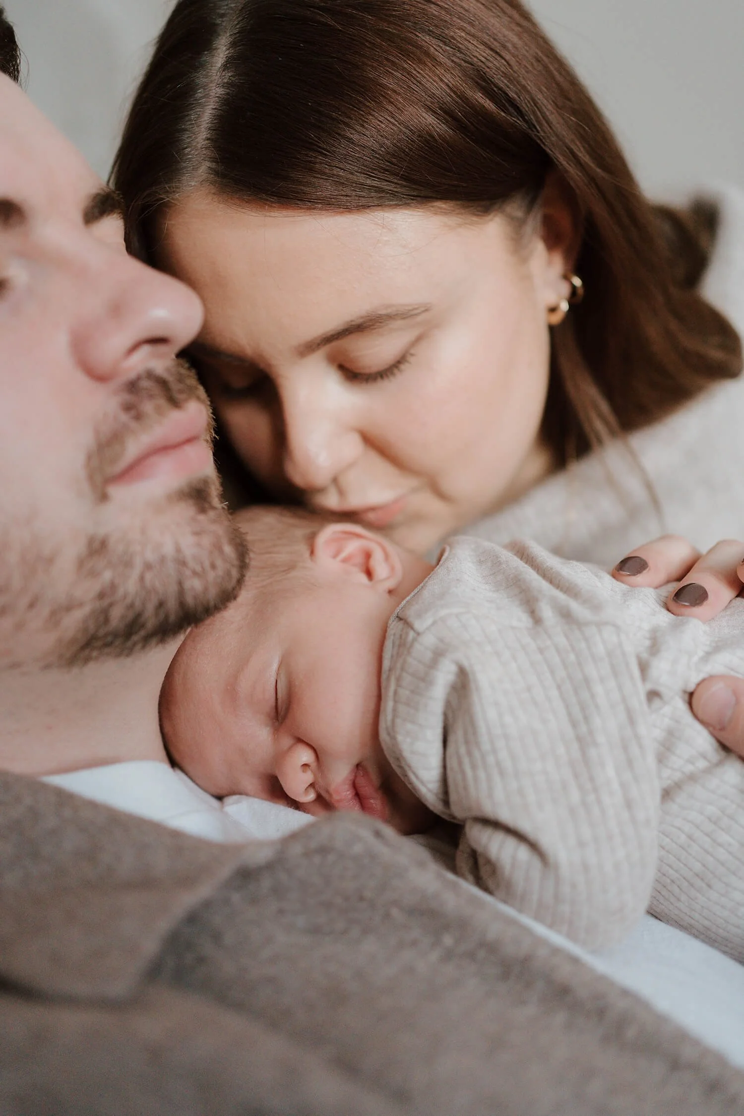 A family of three, a man, a woman, and a newborn baby, lying close together with the baby sleeping on the man's chest, and the woman gently kissing the baby's head.