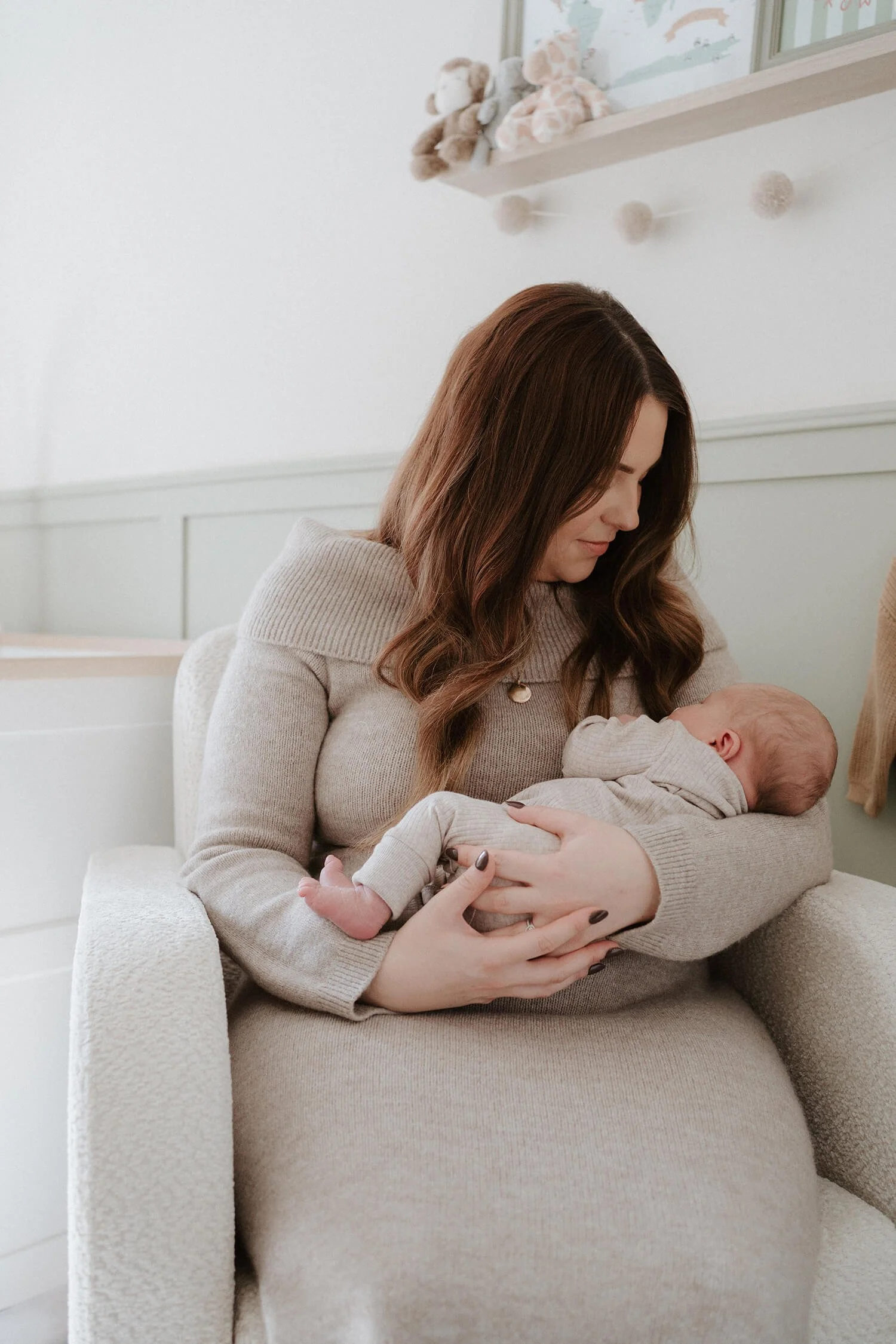 A woman holding a newborn baby while sitting in a beige armchair in a cozy, softly lit room with a World map and stuffed toys on the shelf above.