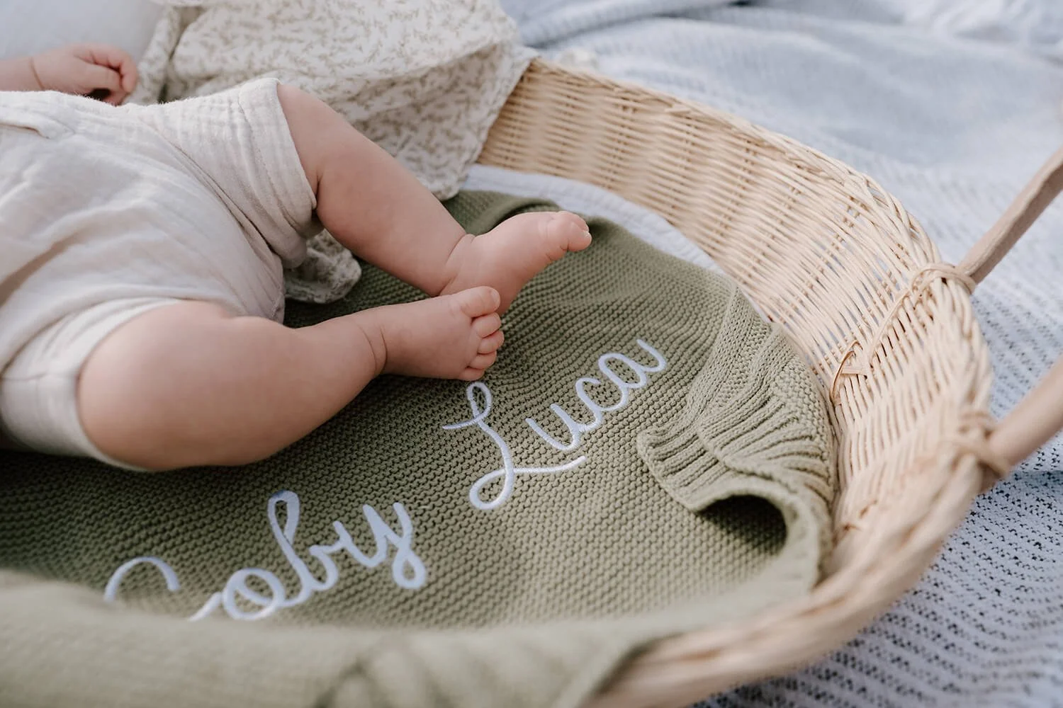 Close-up of a baby's leg and foot lying in a wicker basket with a green knit blanket that has white embroidered text reading 'hello soul'