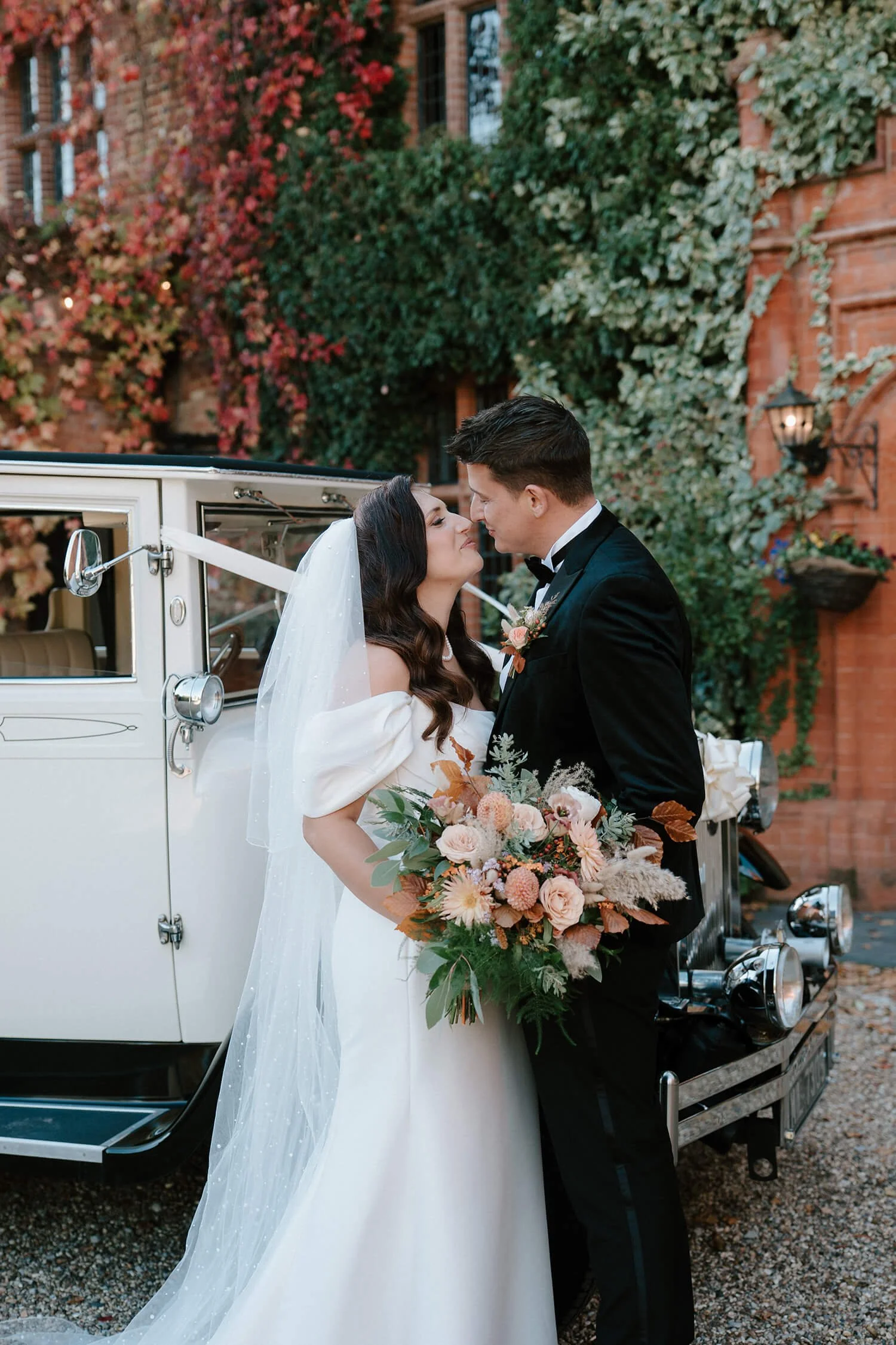 Bride and groom in wedding attire sharing a romantic moment in front of a vintage white car, with brick wall and greenery in the background.
