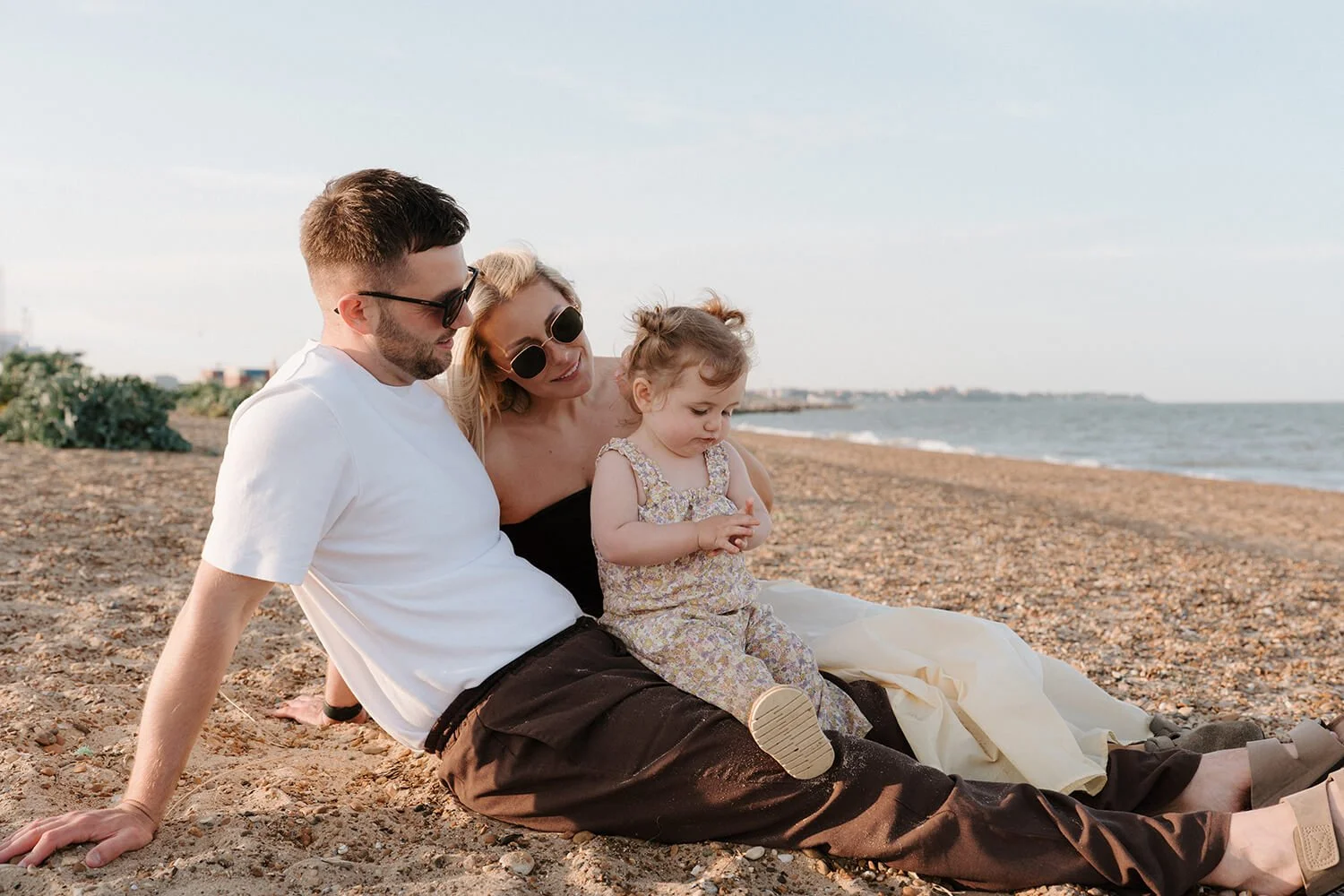 A family of three sitting on a pebble beach near the ocean, with the parents and young girl looking at something on the ground.