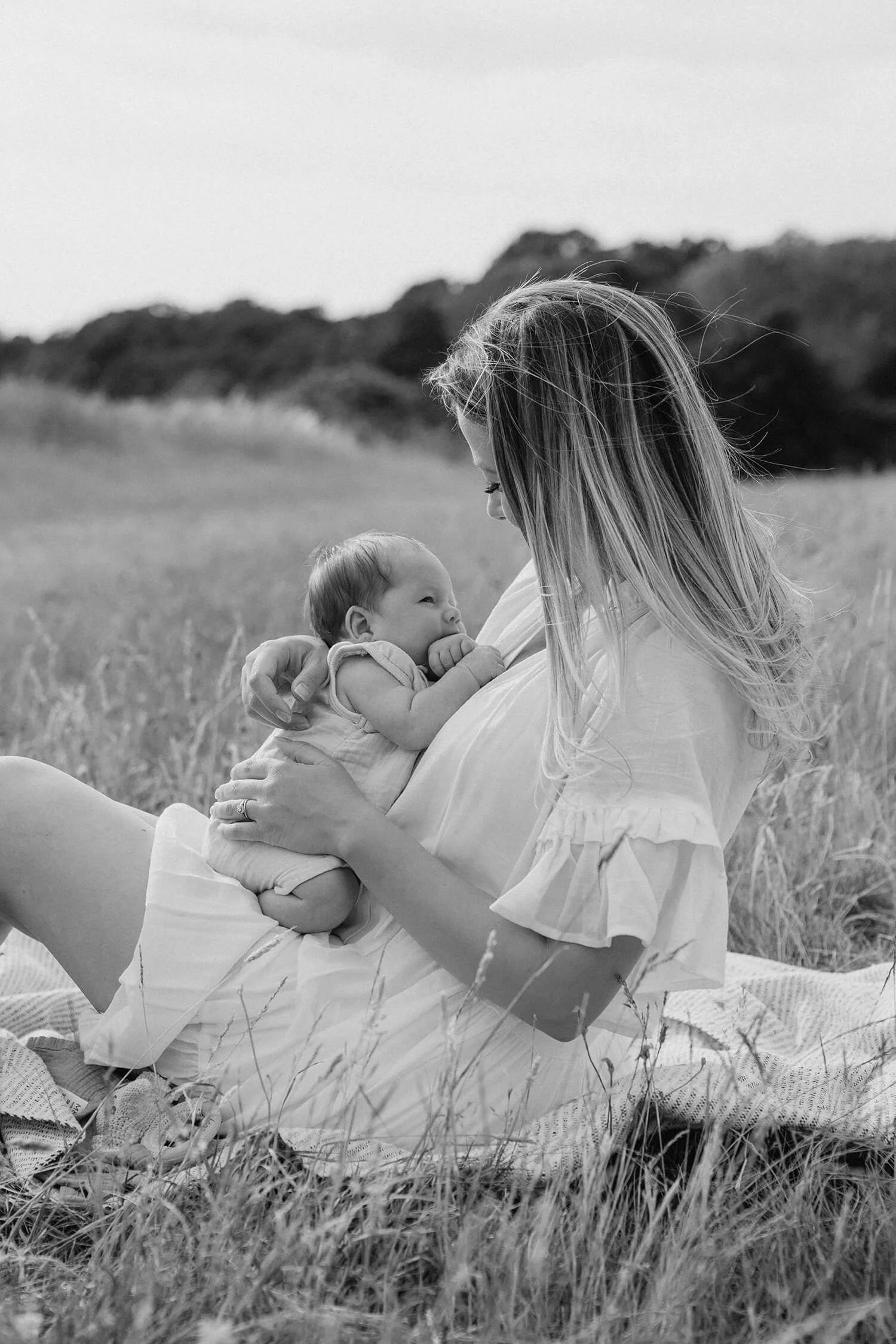 A woman sitting on grass outdoors holding a newborn baby, both facing each other, in black and white.