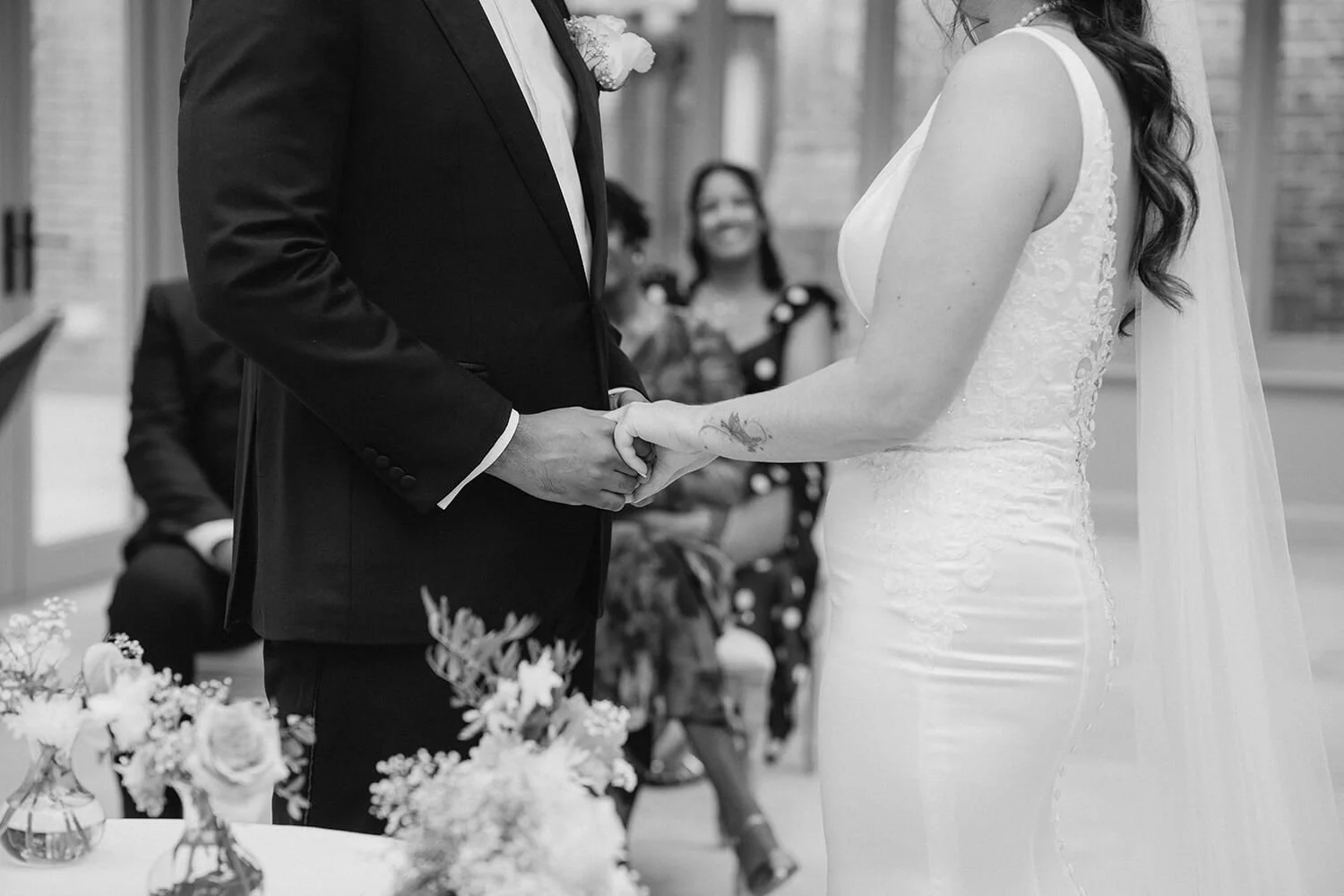 A couple holding hands during a wedding ceremony, with a bride wearing a lace wedding gown and a groom in a black suit. In the background, guests are seated and smiling.