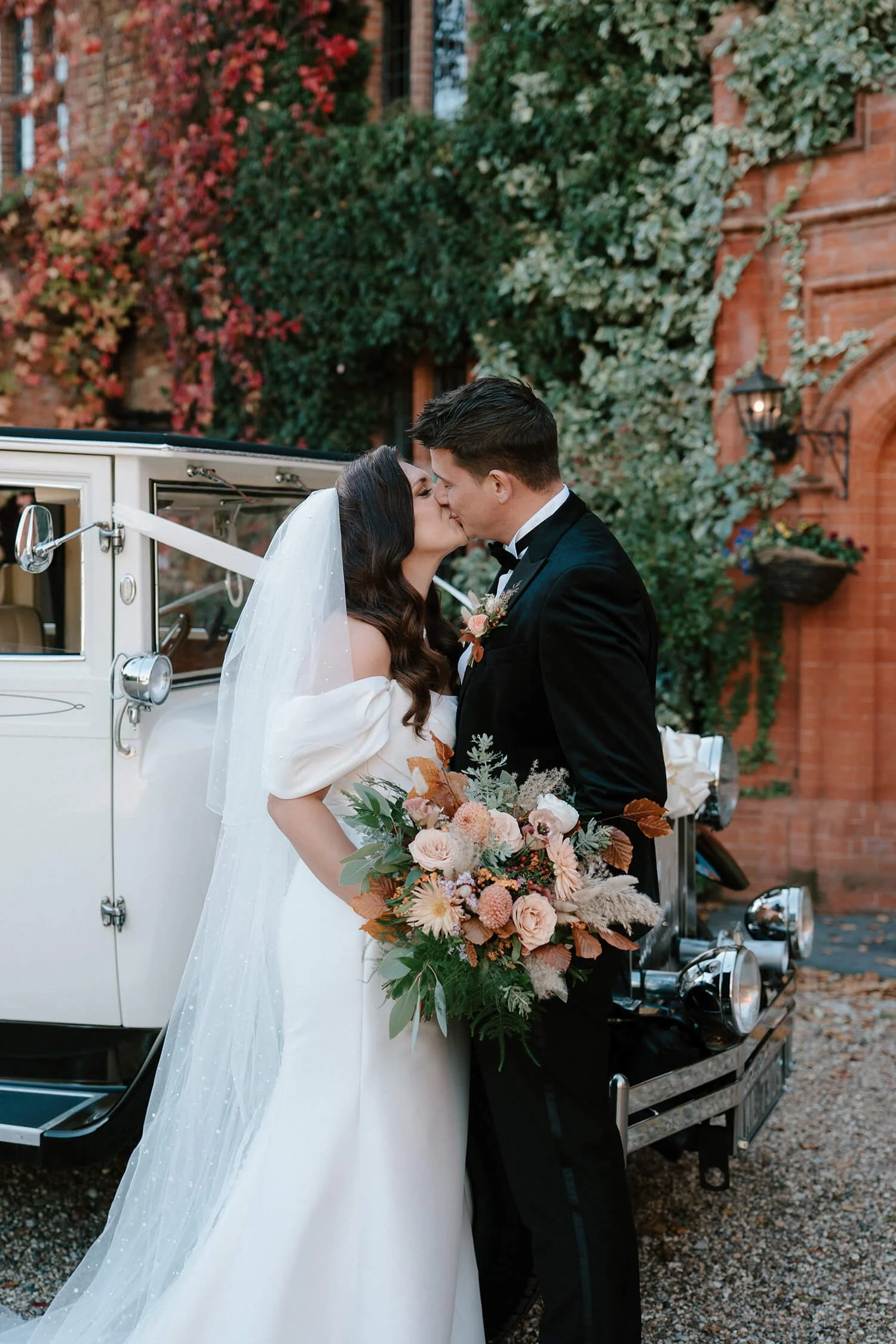 A newlywed couple sharing a kiss in front of a vintage car with a bouquet of flowers, outdoors against a backdrop of brick walls and ivy.