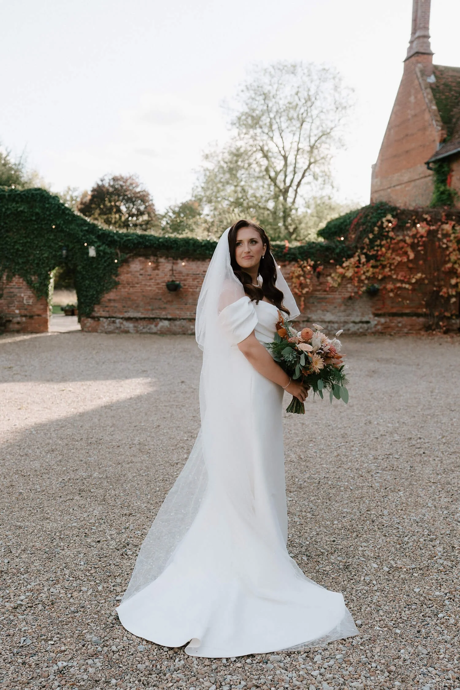 Bride in white wedding dress holding a bouquet of flowers in an outdoor setting with brick walls and trees.