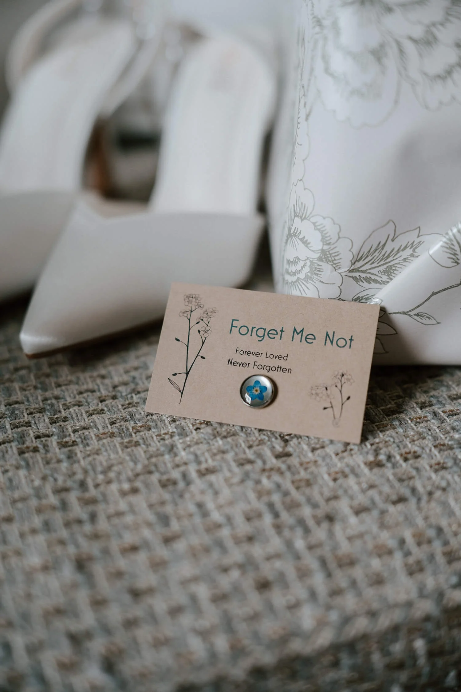 A small card with the words 'Forget Me Not,' 'Forever Loved,' and 'Never Forgotten,' decorated with a floral illustration, placed on a textured fabric surface. A pair of white high-heeled shoes and a white cloth with floral patterns are partially vis