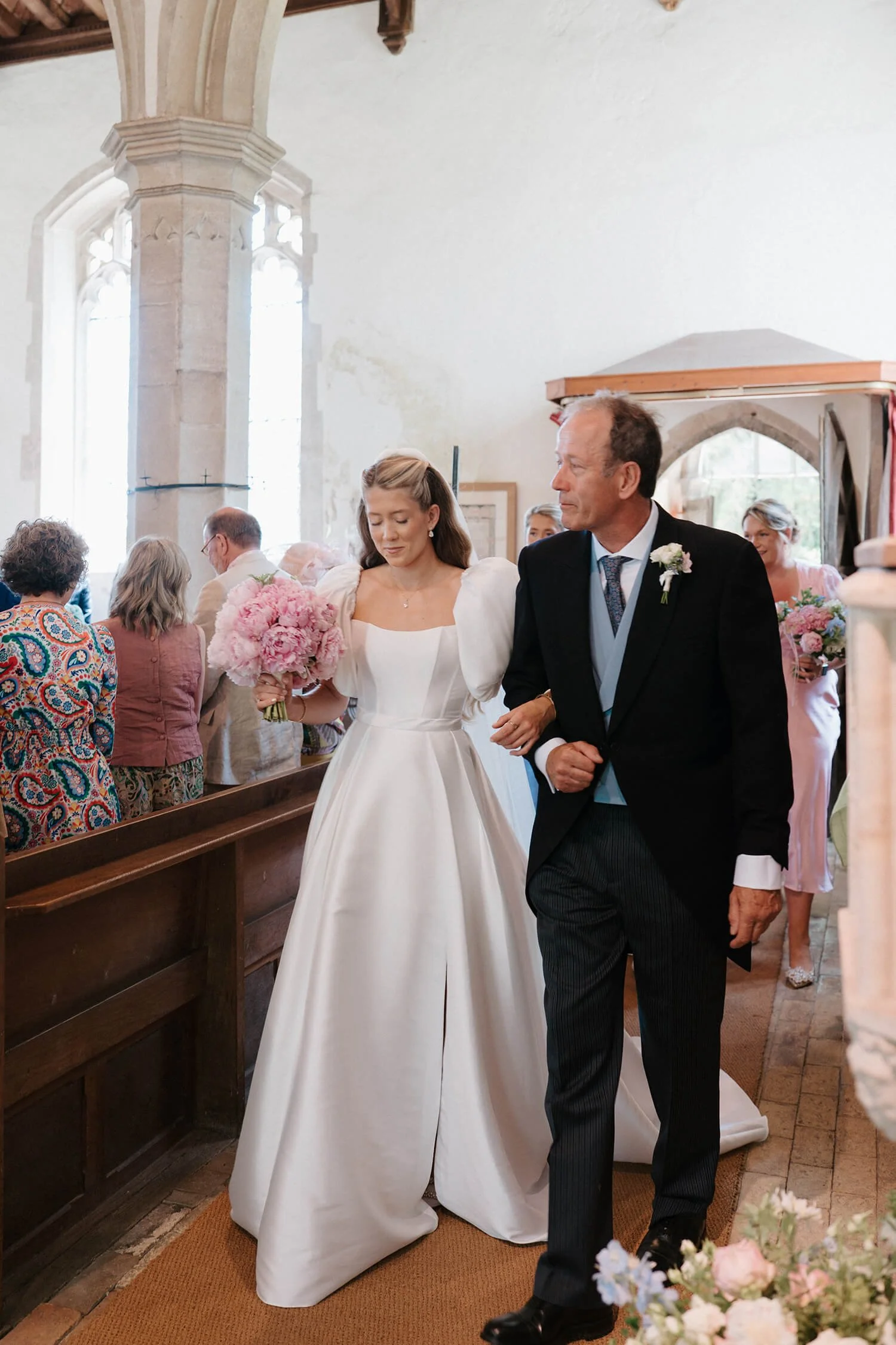 A bride in a white wedding dress holding a bouquet, walking down the aisle with an older man, possibly her father, at a church wedding ceremony.