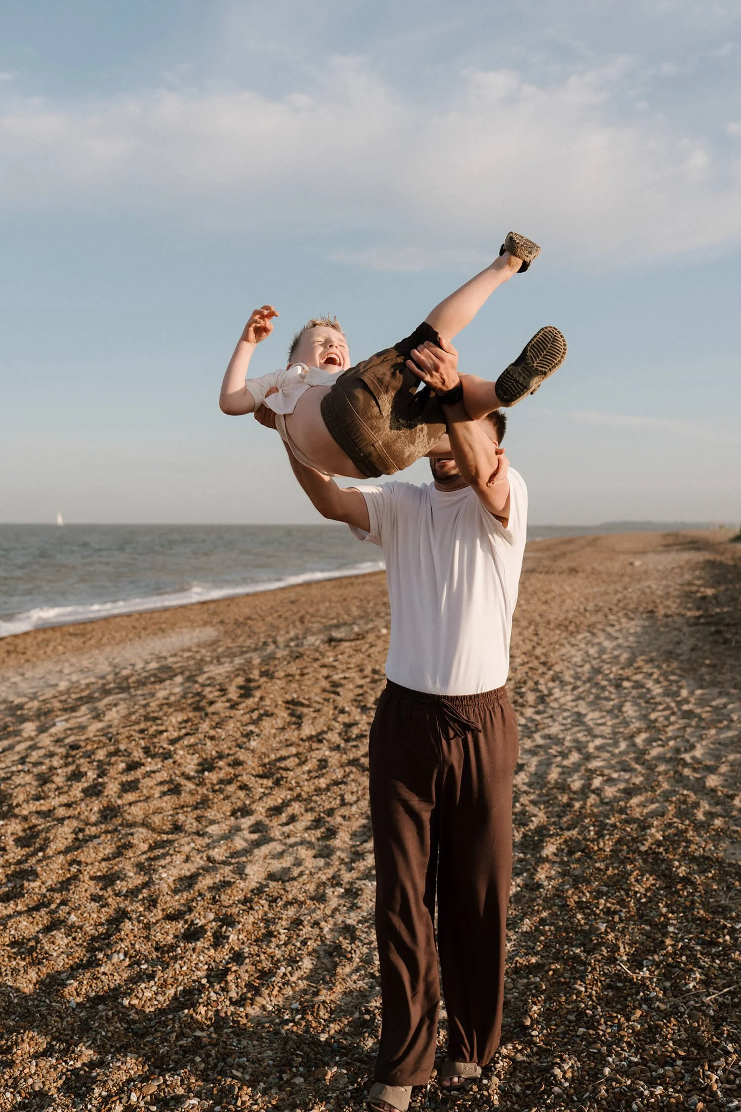 A man lifting a young boy in the air on a beach with the ocean in the background.