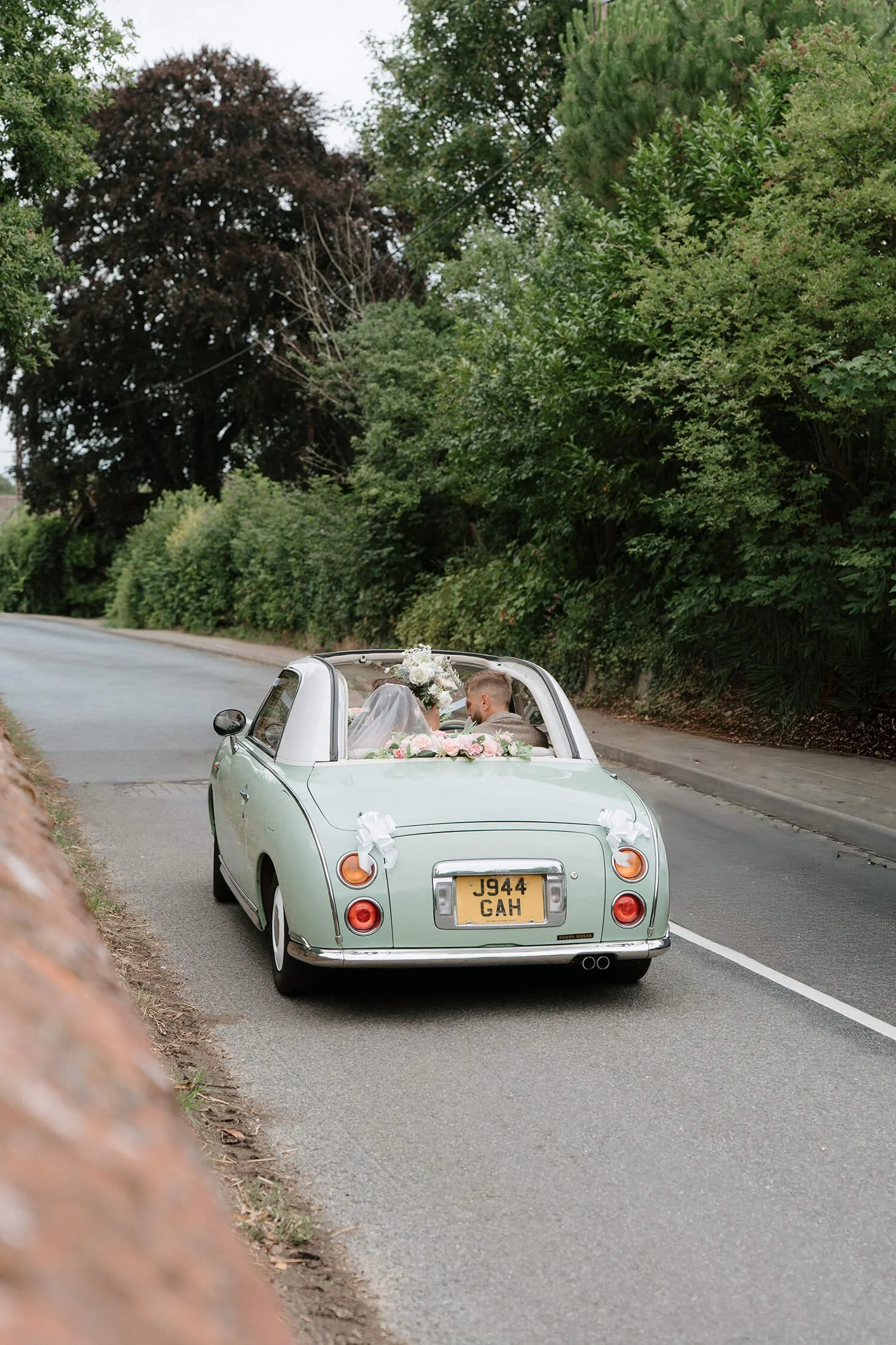 Vintage light green convertible car decorated with flowers, driving on a rural road surrounded by lush green trees, with a bride and groom inside celebrating their wedding.