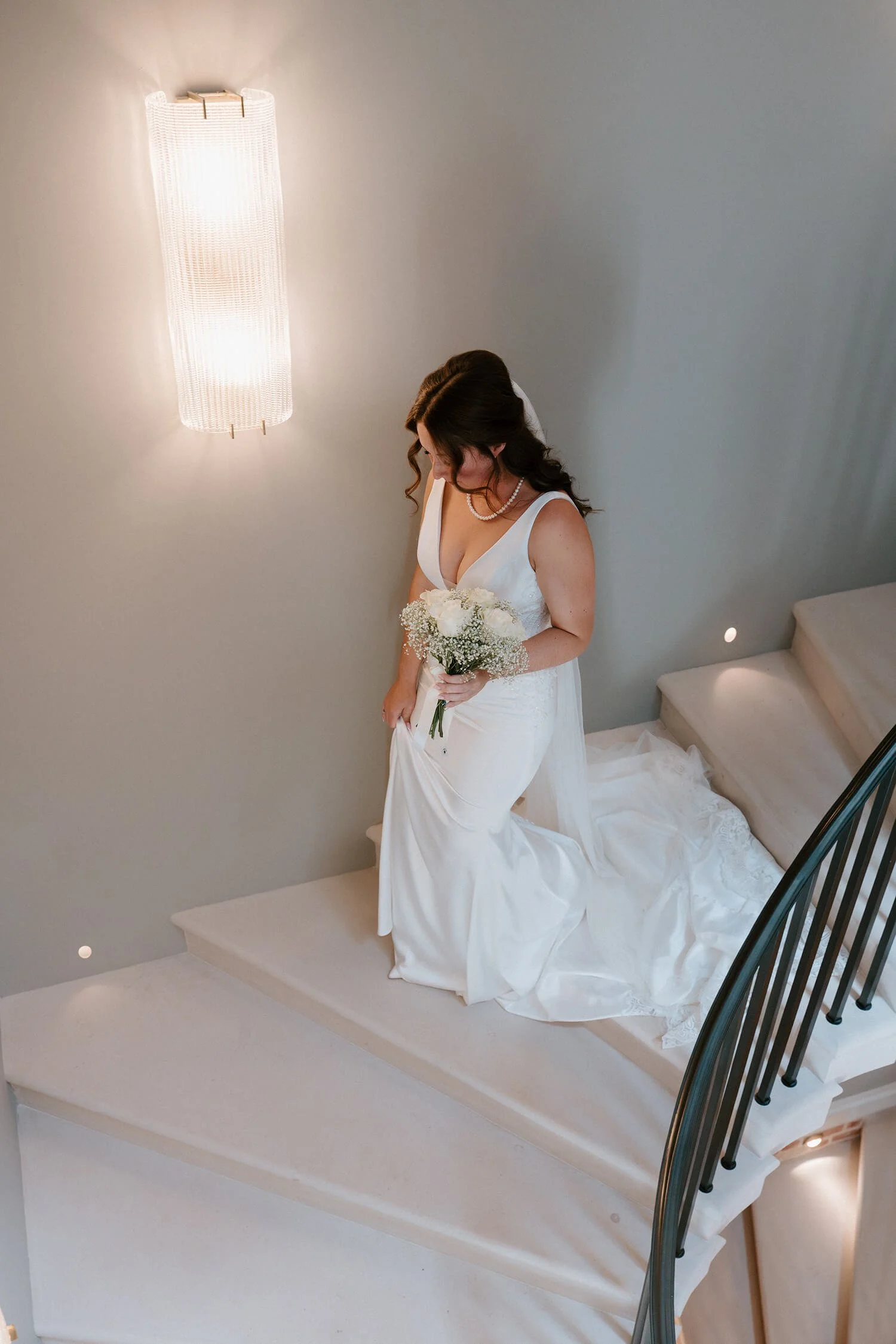 A bride in a white wedding gown holding a bouquet of white flowers, standing on a staircase in a minimalist, softly lit interior.