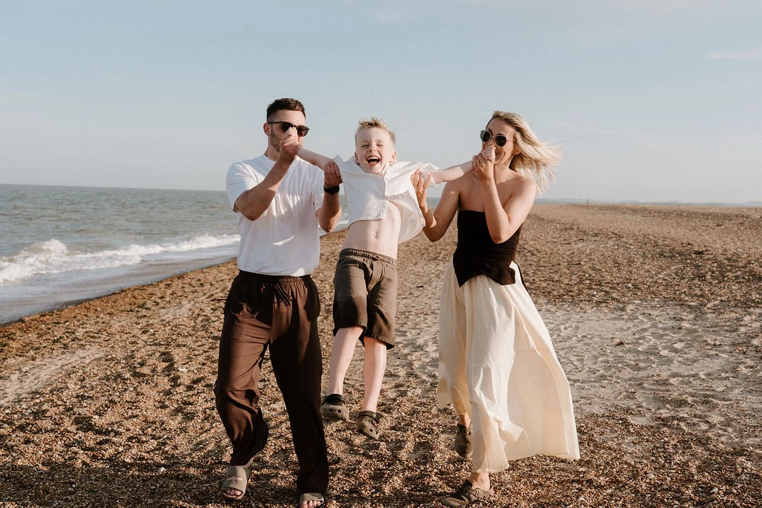 A family of three on a beach, with a man and woman holding a young boy who is smiling and laughing, all dressed casually and enjoying a sunny day by the ocean.