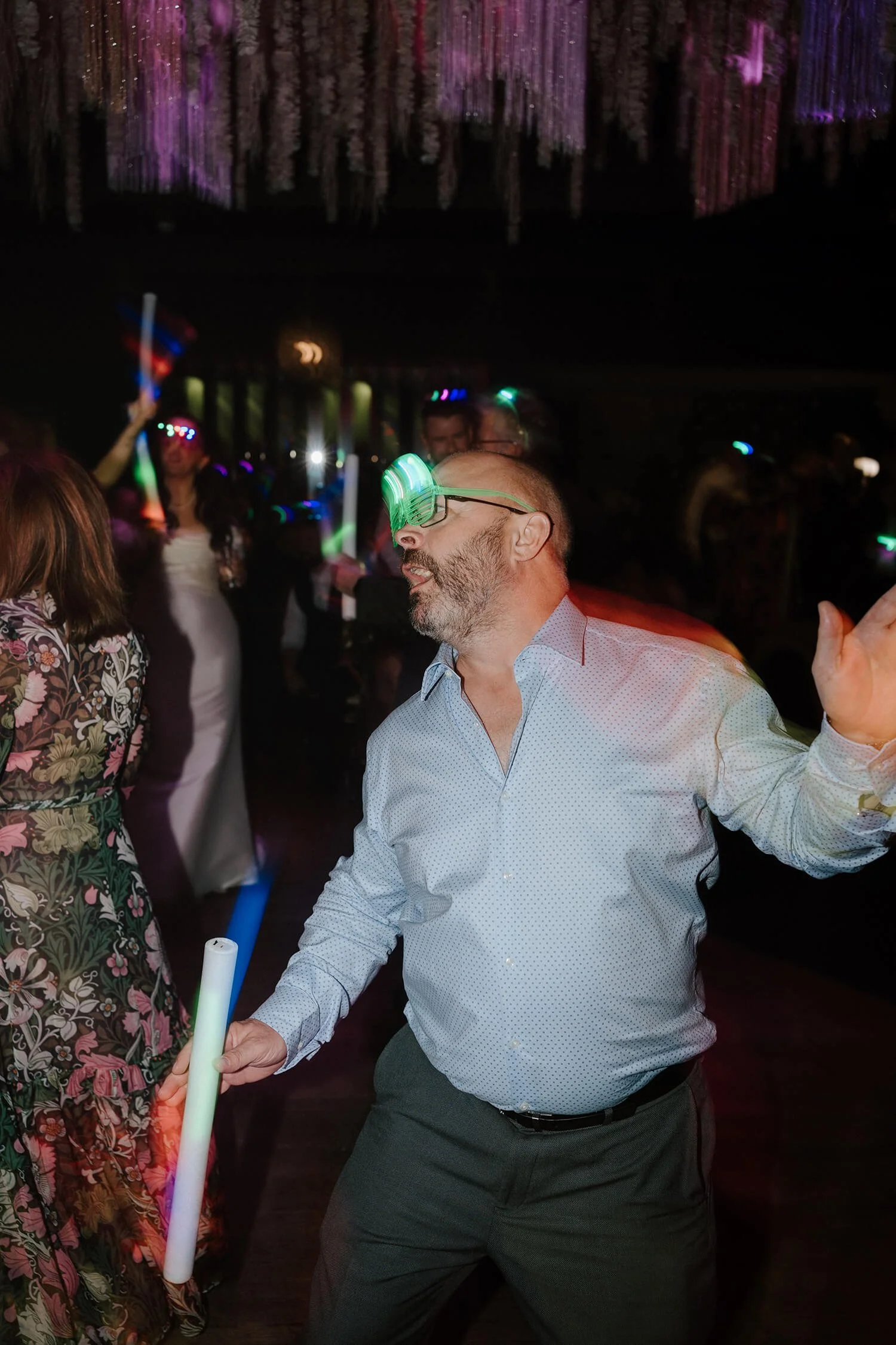 Man dancing at a party wearing glow glasses, surrounded by other partygoers with glow sticks and colorful lights.