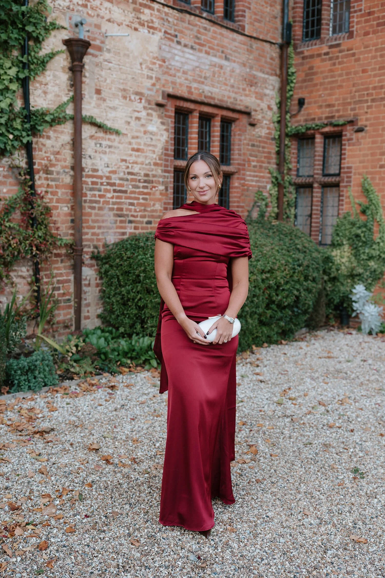 Young woman in a long, elegant, off-the-shoulder red gown holding a white clutch bag, standing outdoors in front of a brick building with greenery.