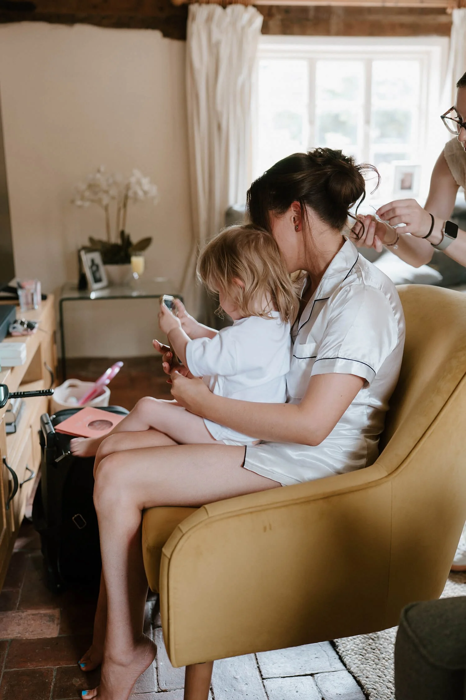 A woman sitting on a mustard yellow armchair, with a young girl sitting on her lap. The woman is getting her hair styled by a second person, and the girl is looking at a phone. They are in a cozy, well-lit room with a window behind them, and a dresse