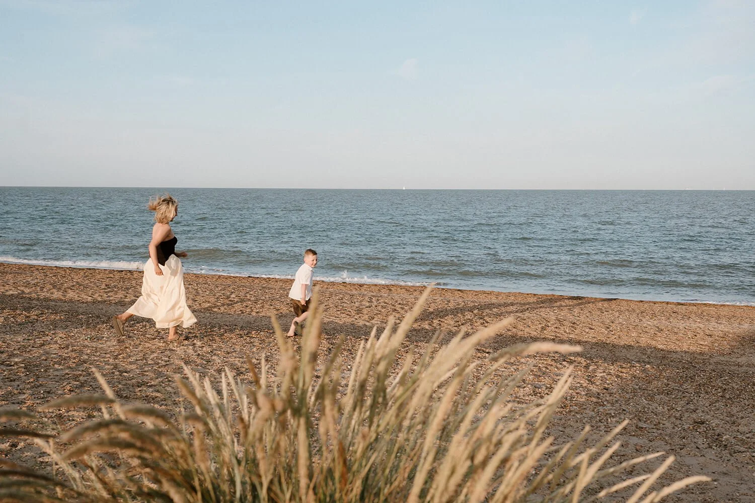 A woman and a young boy running along a sandy beach near the ocean, with beach grass in the foreground and the horizon in the background.