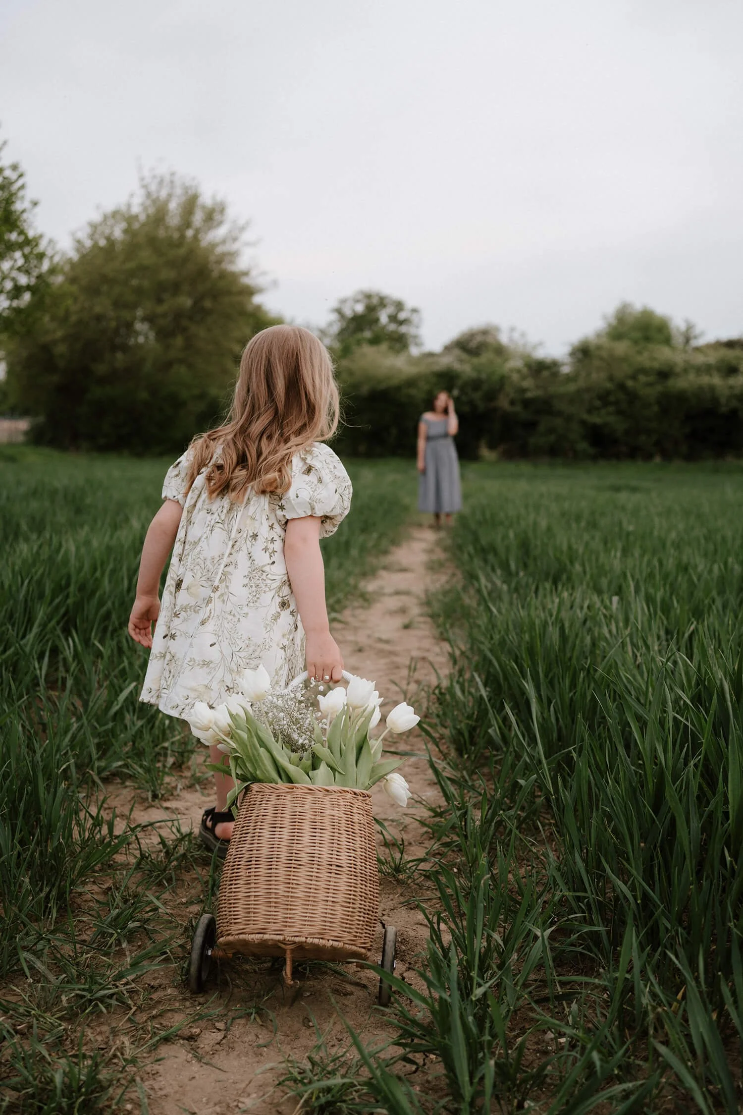 A young girl with red hair in a floral dress is walking along a dirt path in a green field, holding a wicker wagon filled with white tulips. In the background, a woman in a gray dress stands amidst trees.