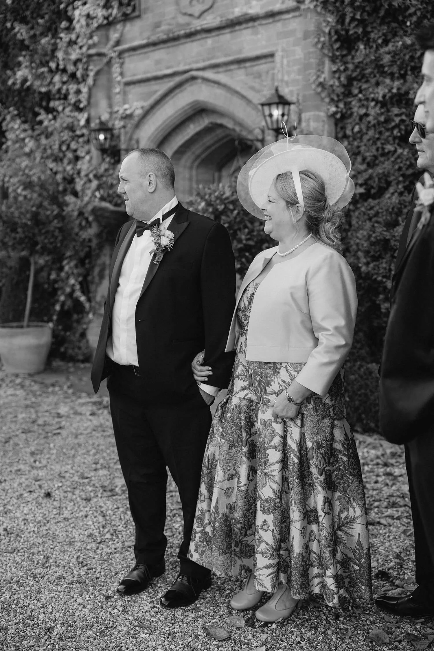 Black and white photo of a couple, the man in a tuxedo and the woman in a floral dress and light jacket, standing outdoors in front of a stone building with ivy, smiling and holding hands. The woman is wearing a large hat with a veil, and there is a 