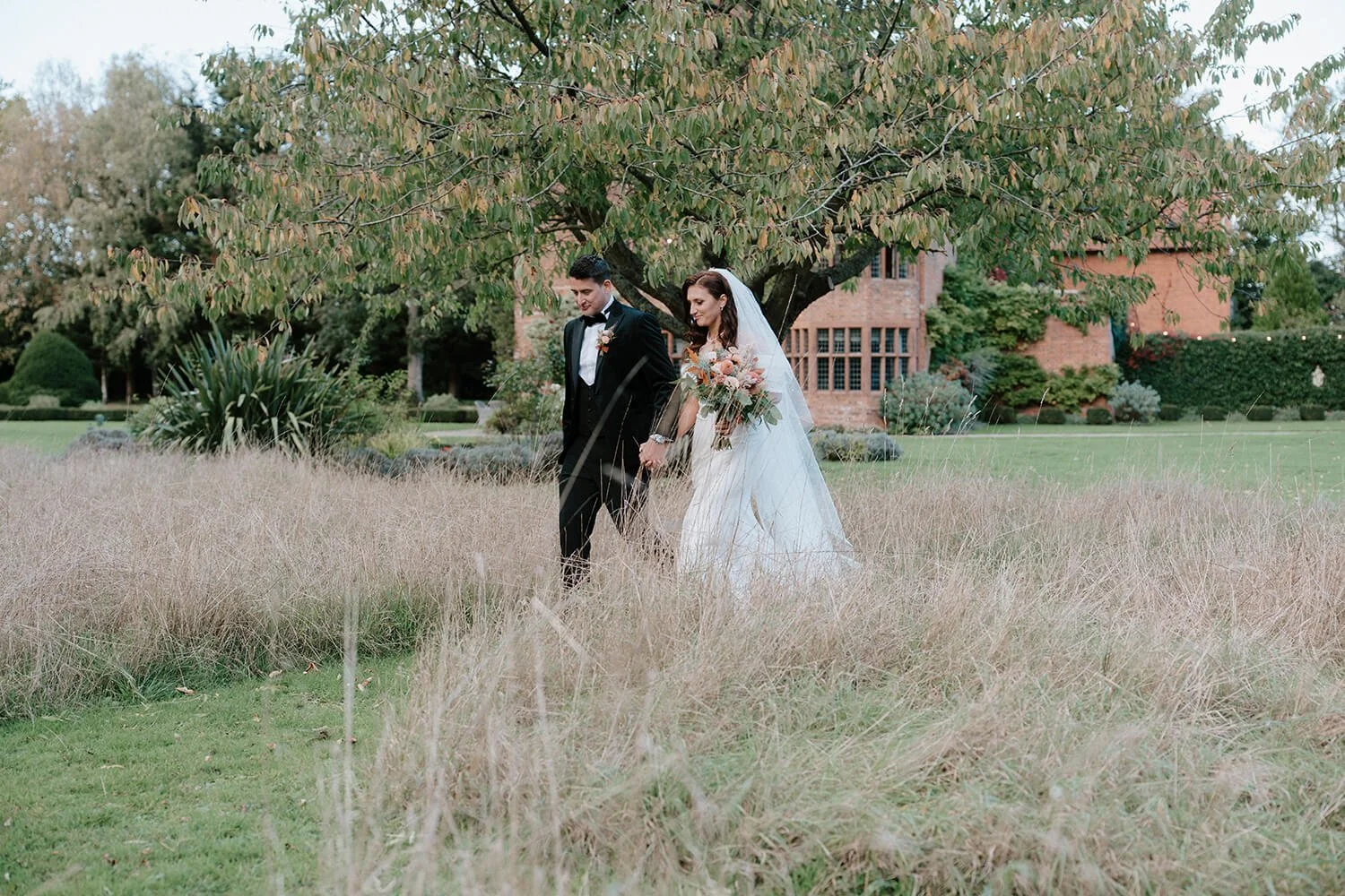 A bride and groom walking hand in hand through a grassy field, with a large tree and a brick building in the background.