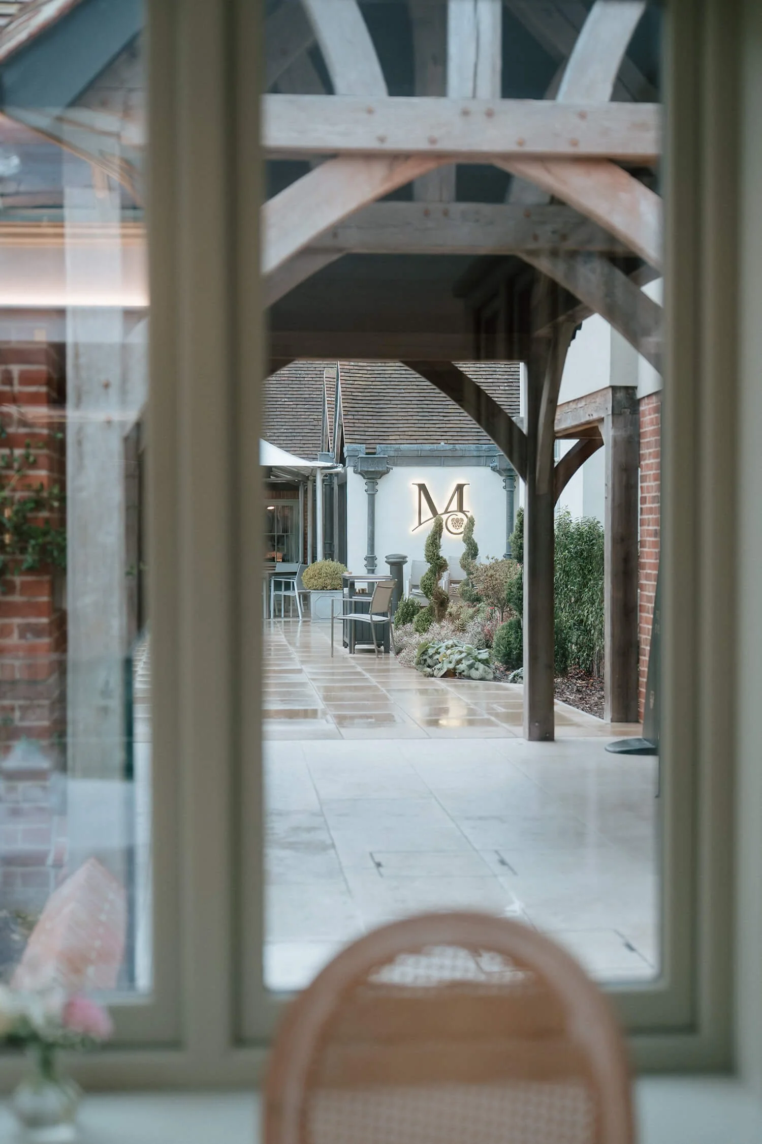 View through a window showing an outdoor terrace with wooden beams, potted plants, and a lit sign with a large letter 'M' on a white wall.