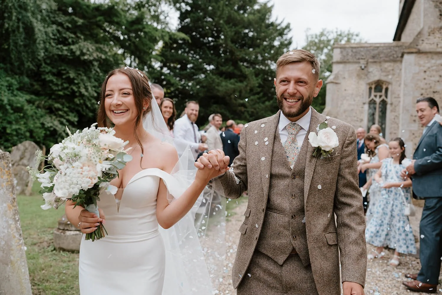 A bride and groom smiling and holding hands after their wedding ceremony, surrounded by friends and family outside near an old stone building, with confetti falling around them.