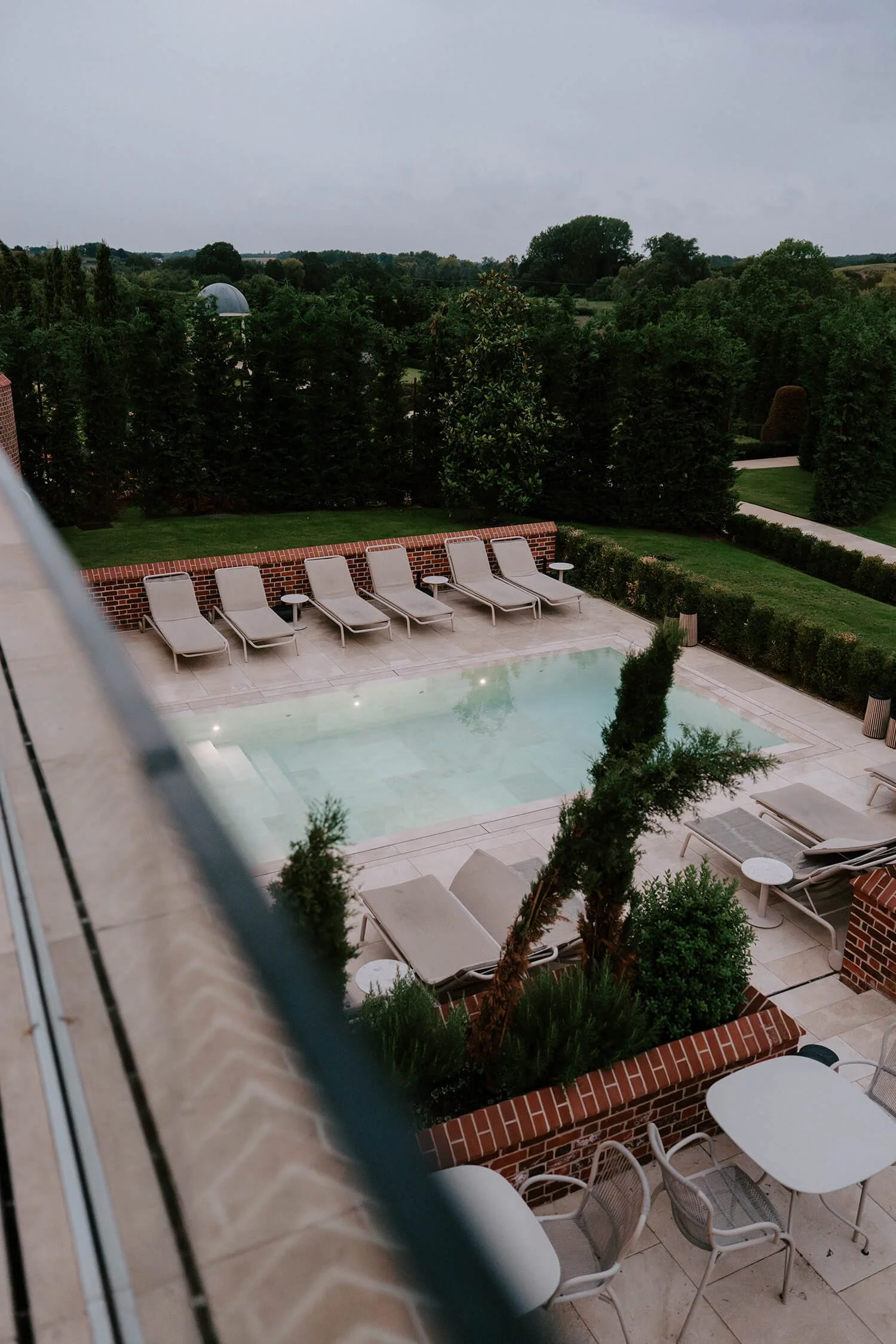 View of a backyard swimming pool surrounded by lounge chairs, small tables, and lush greenery, with a cloudy sky overhead.