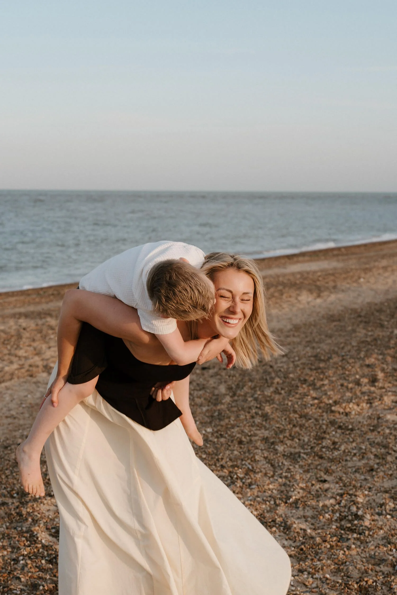 A woman giving a piggyback ride to a young boy on a beach with the ocean in the background, both smiling and enjoying the moment.
