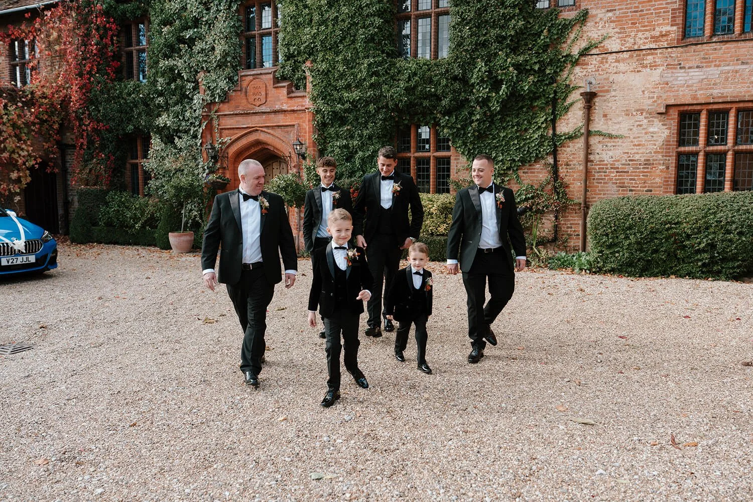 Group of men and boys dressed in tuxedos walking outside in front of a brick building with ivy and windows.