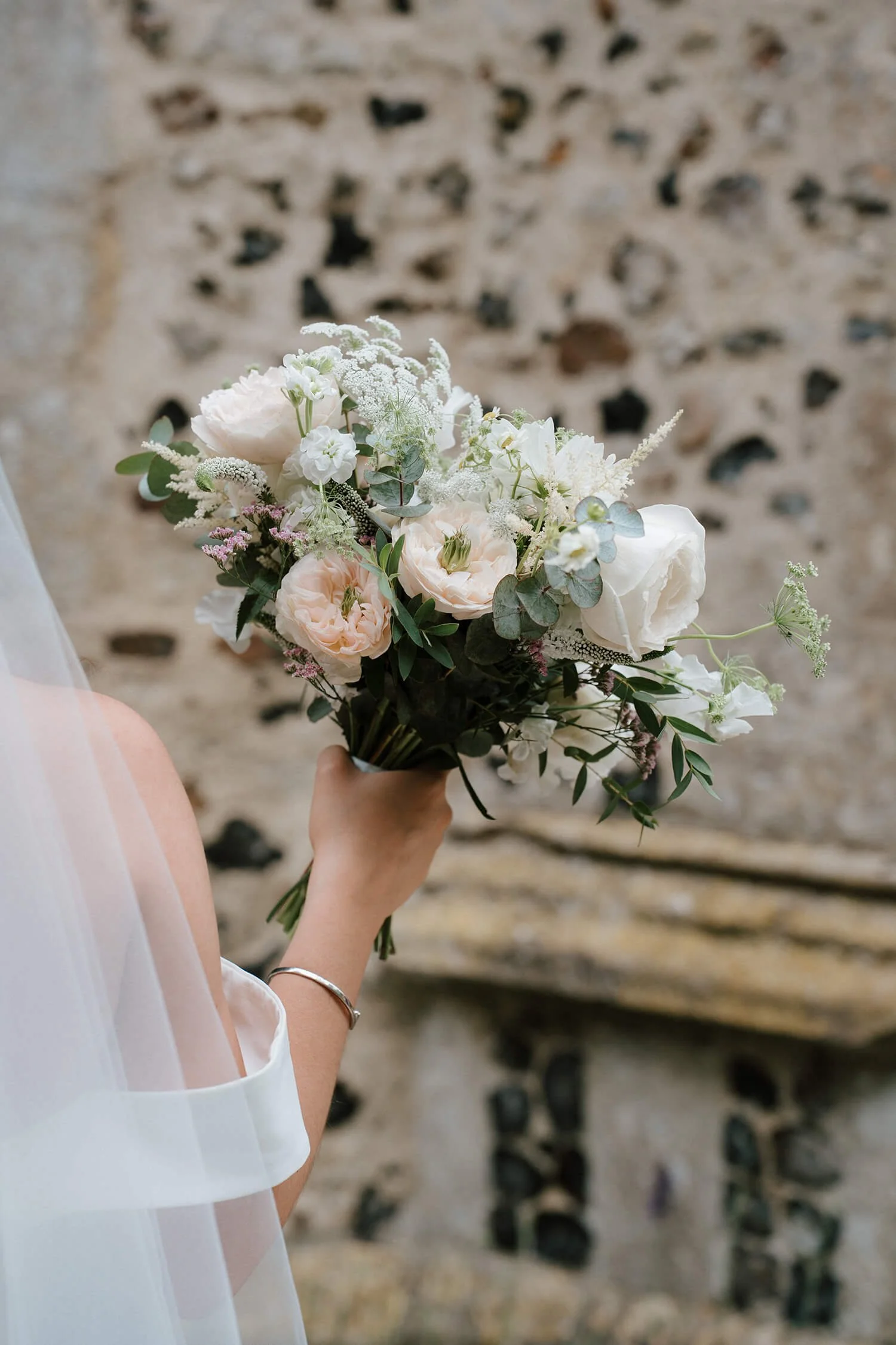 A person holding a bouquet of white and pastel flowers, with a stone wall in the background.