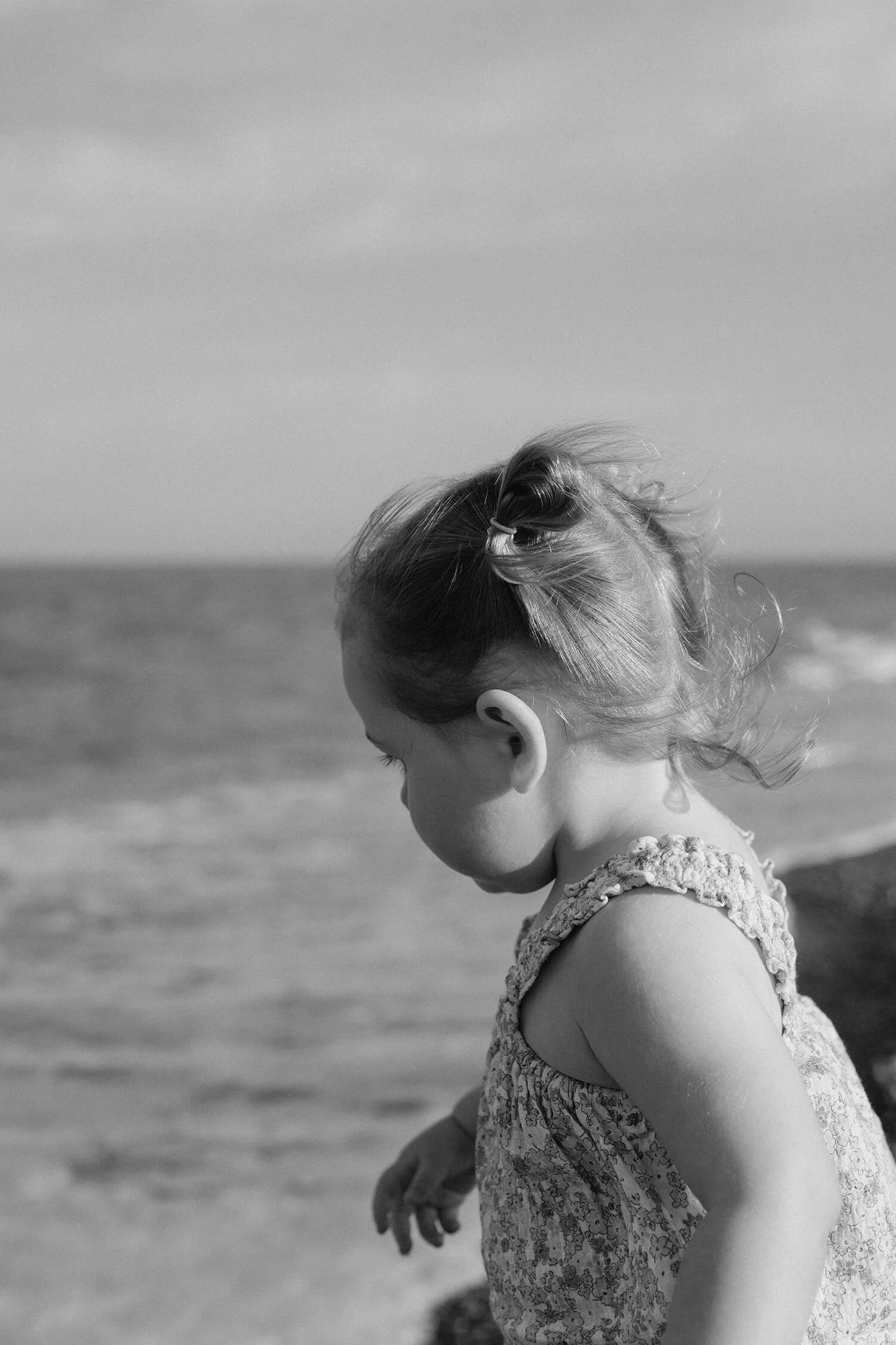 A young girl with her hair tied up in a small ponytail, looking down at the ground near the ocean shore, in black and white.