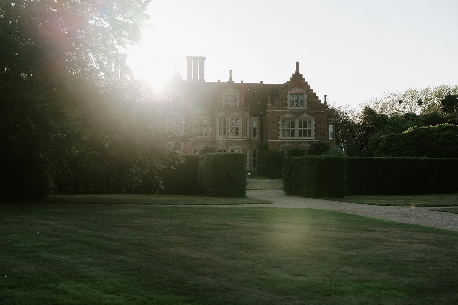 A large, historic brick house with many windows, surrounded by well-maintained bushes and trees, with the sun shining through the trees in the background.
