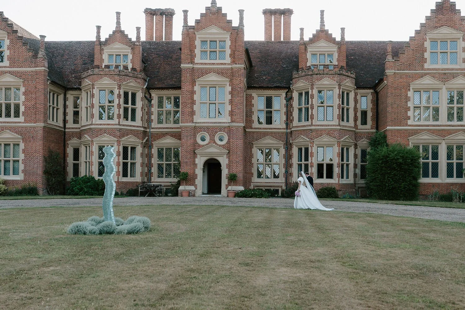 A bride and groom standing on a lawn in front of a large red brick Gothic-style mansion with many windows and chimneys. The bride wears a white wedding dress and veil, while the groom is in a dark suit. There is a modern sculpture on the lawn to the 