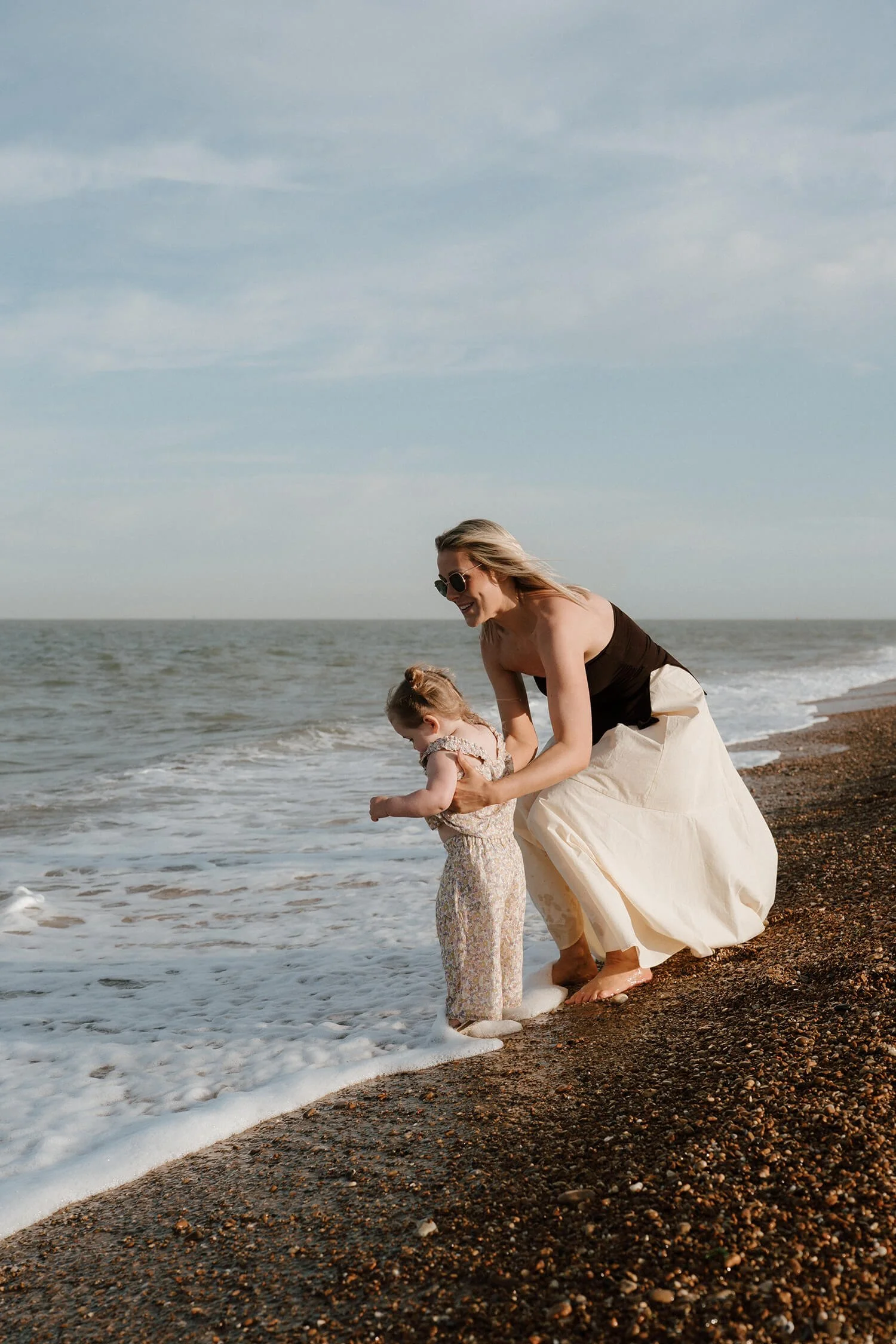 A woman and a young girl on a beach, playing by the surf with the sky in the background.