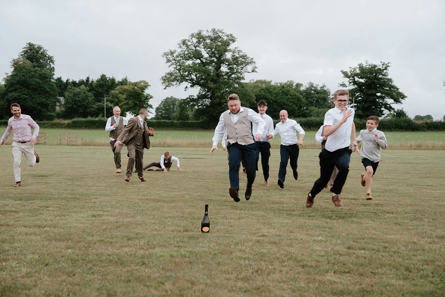 Group of men and boys in formal and casual attire running and playing with a bottle on an open grassy field with trees in the background.