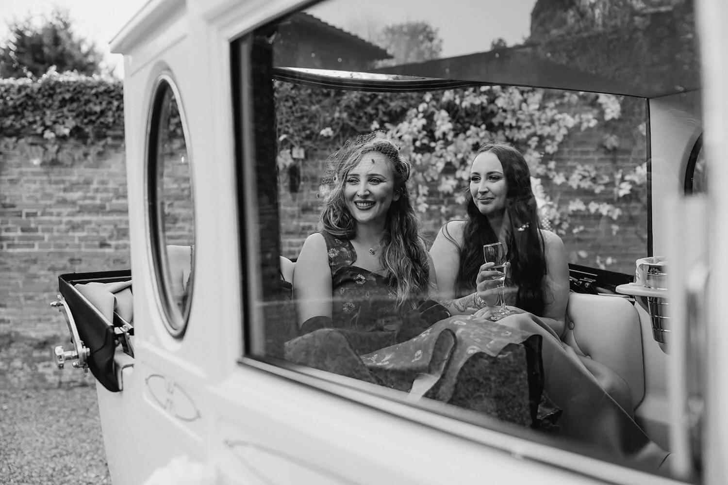 Two women sitting in a vintage car, one holding a champagne glass, smiling and enjoying a celebration outdoors in a garden with a brick wall and flowering bushes in the background.